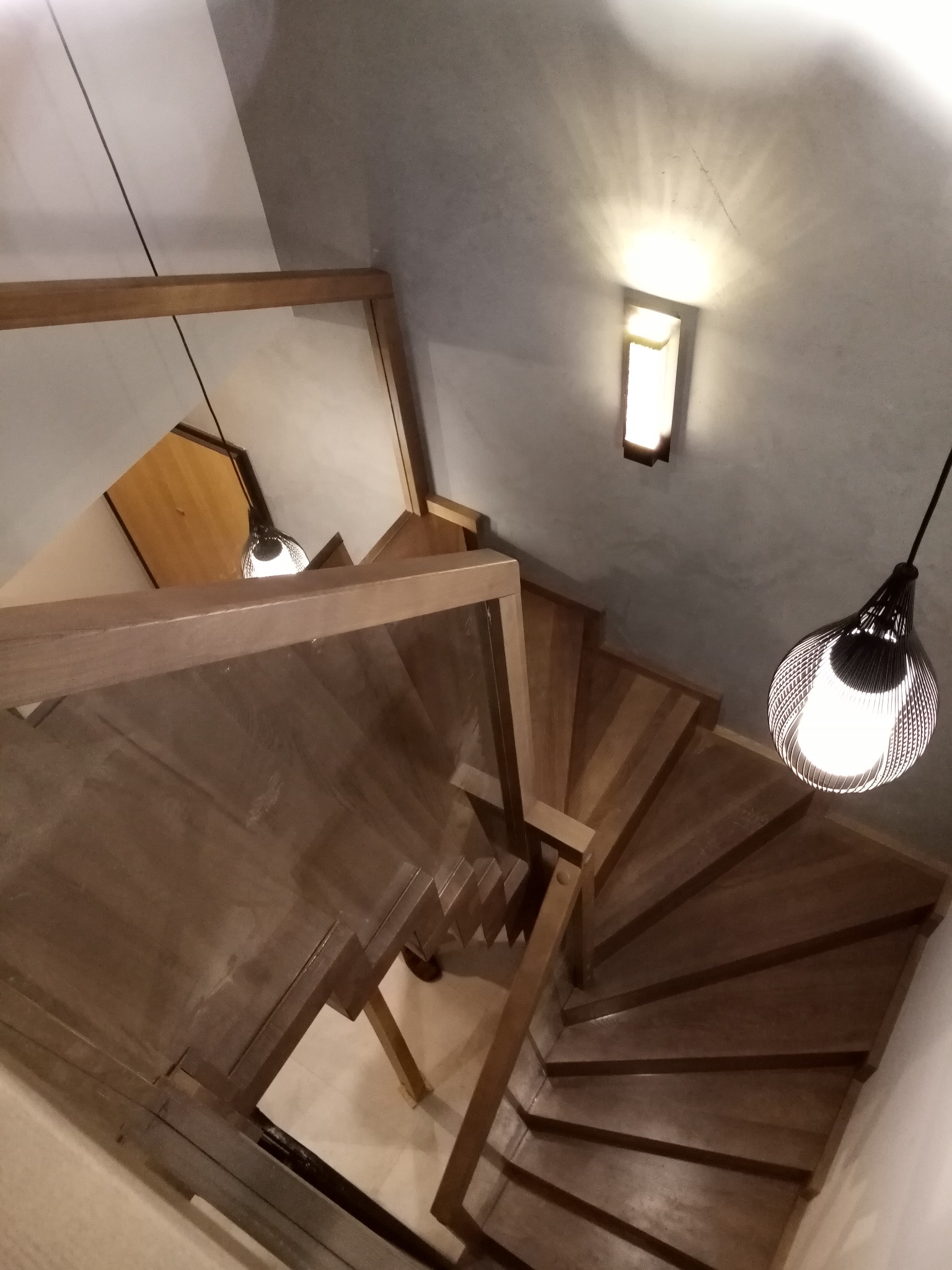 Spiralne stopnie z jasnego drewna widziane z góry, oświetlone wiszącą lampą z ażurowym kloszem i kinkietem na szarej ścianie.