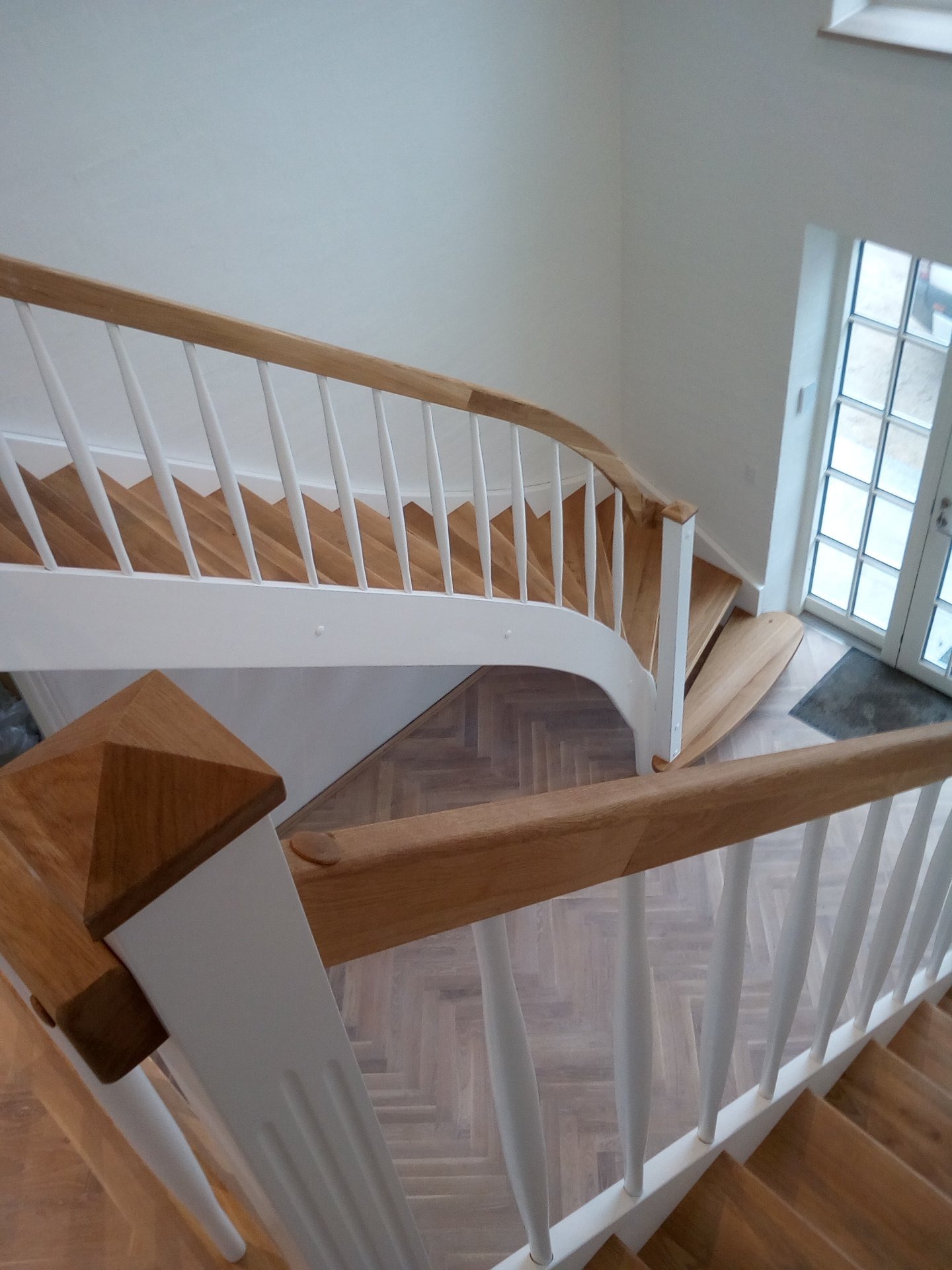 Drewniane schody z białymi balustradami i poręczą z jasnego drewna, widok z góry na parkiet w jodełkę i przeszklone drzwi w tle.