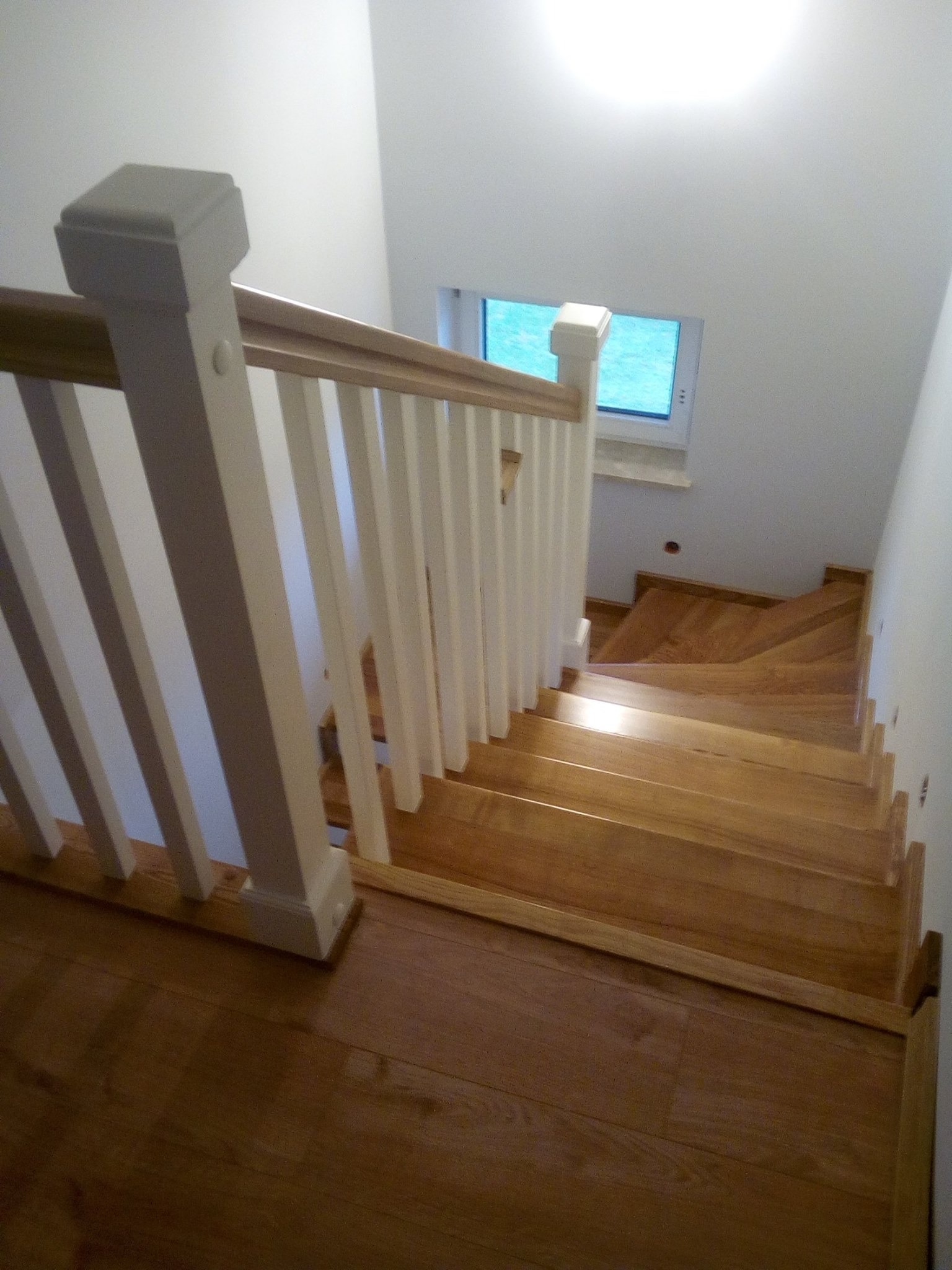Widok z góry na drewniane schody z białą, klasyczną balustradą w jasnym wnętrzu, oświetlone górnym światłem, widoczne małe okno w ścianie.