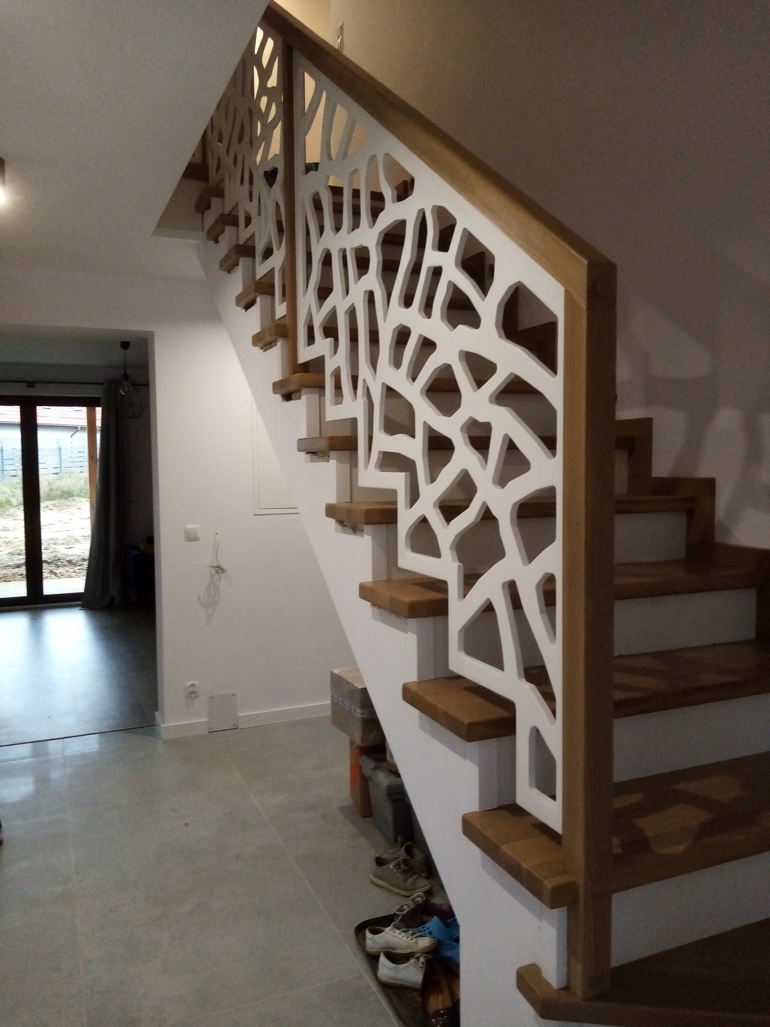 Nowoczesne schody drewniane z białą, ażurową balustradą o organicznym wzorze, widoczne z dołu, z fragmentem podłogi i otwartym przejściem do innego pomieszczenia.