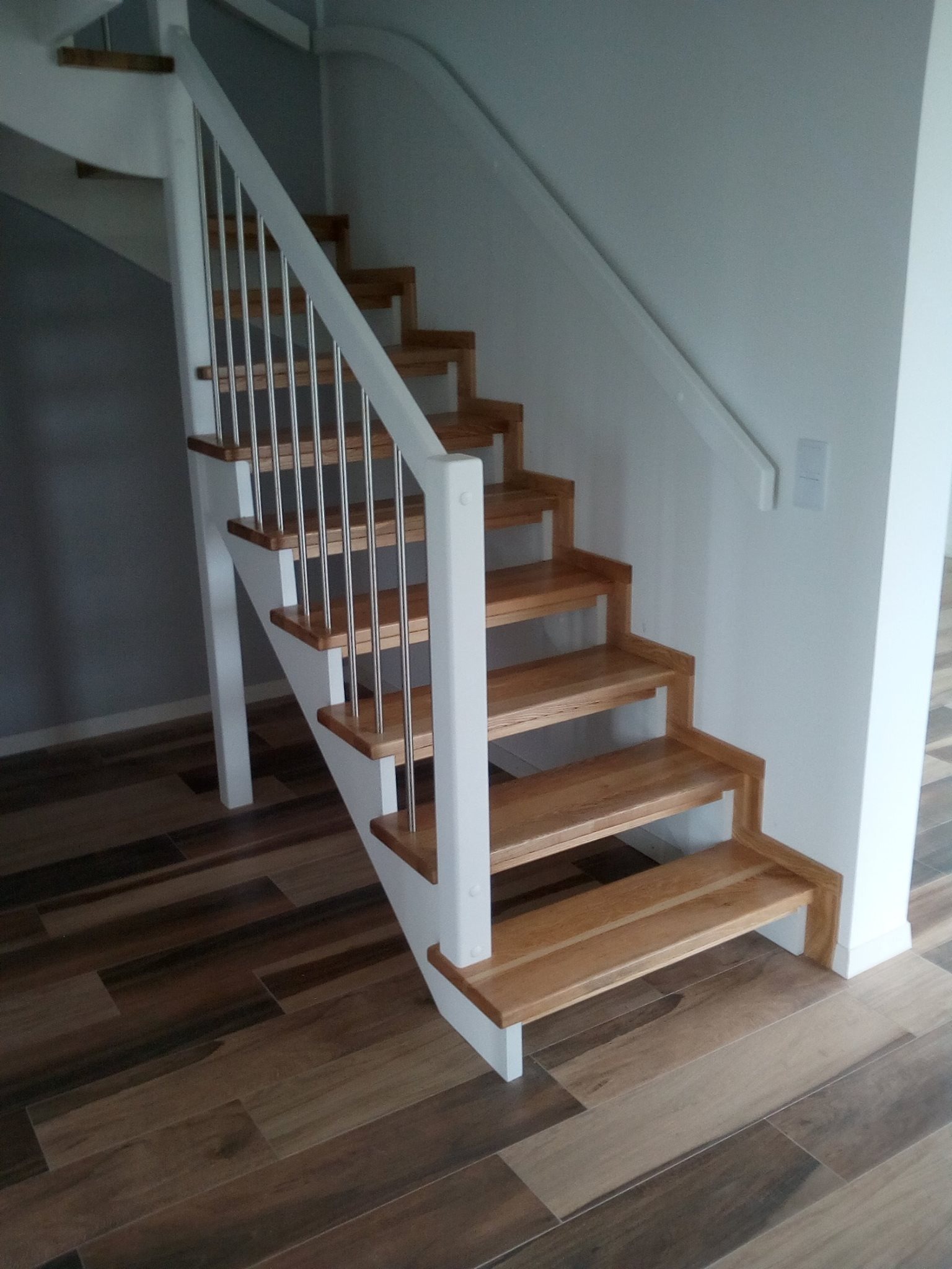 Nowoczesne schody z drewnianymi stopniami i białymi elementami konstrukcyjnymi, uzupełnione metalowymi balustradami, na tle podłogi z płytek imitujących drewno.