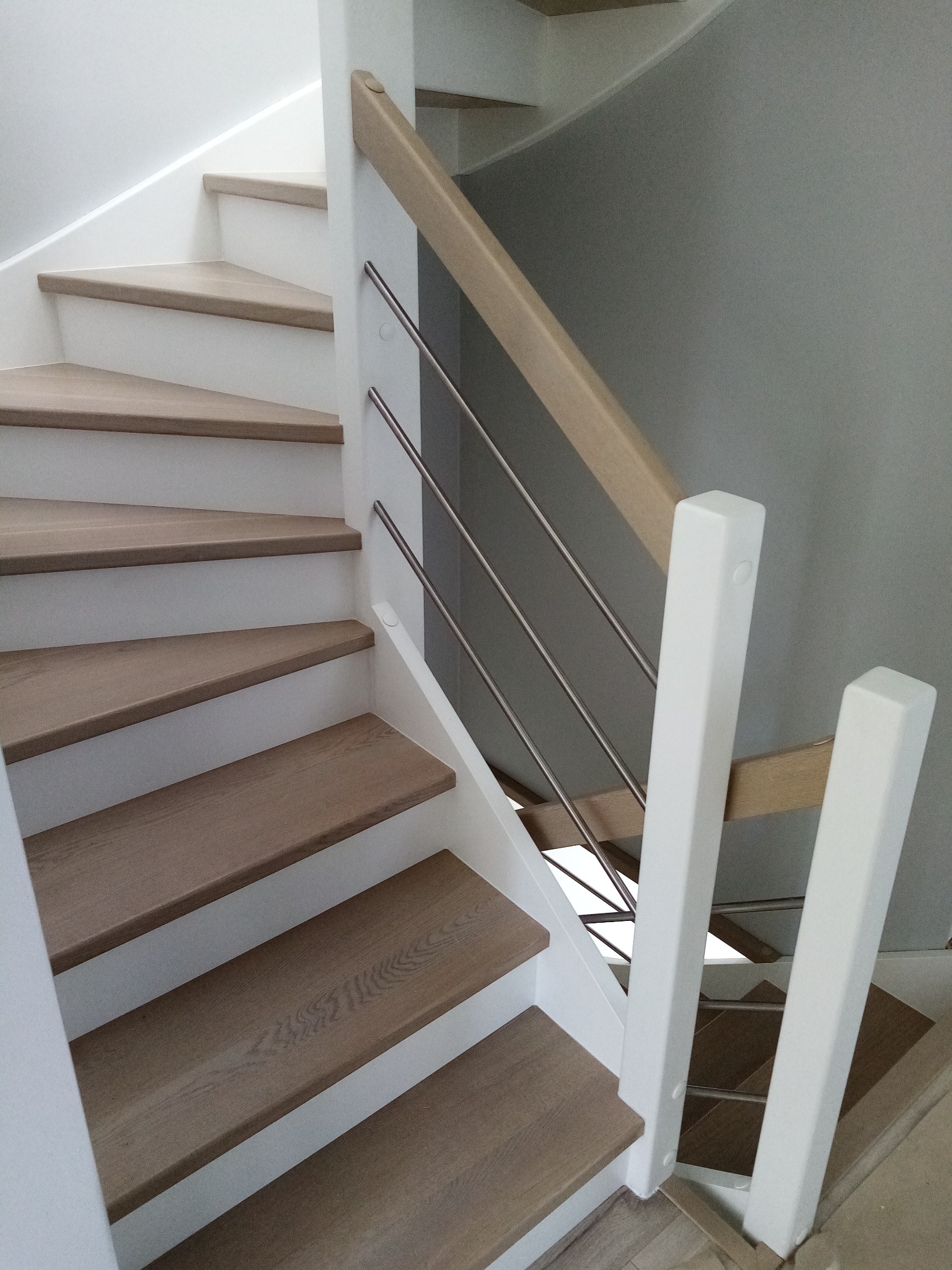 Nowoczesne schody z drewnianymi stopniami i białymi podstopnicami, zabezpieczone poręczą z jasnego drewna i metalowymi prętami.