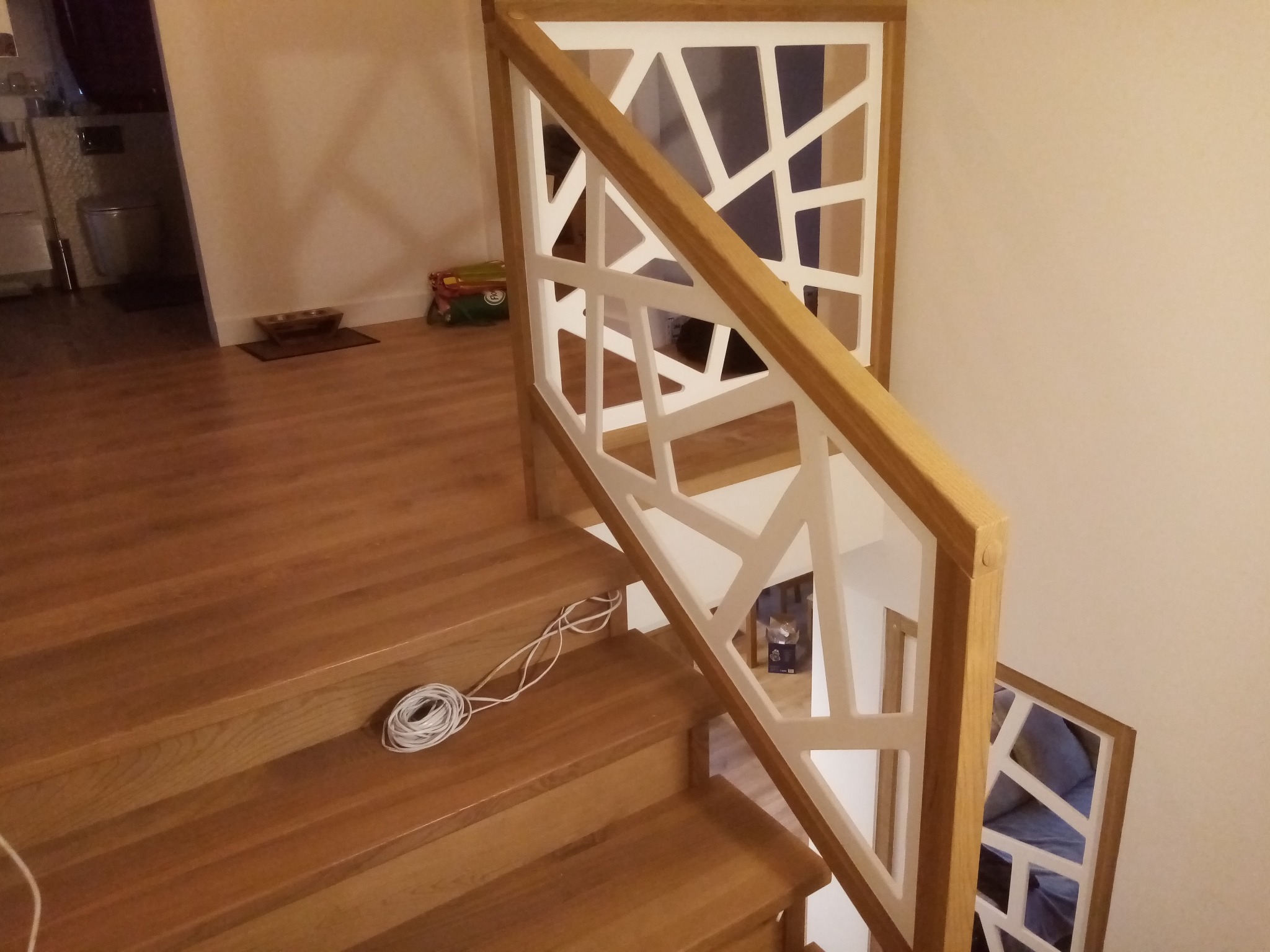 Jasne, drewniane schody z oryginalną, białą balustradą o geometrycznym wzorze, widziane z góry. Na jednym ze stopni leży zwój białego kabla.