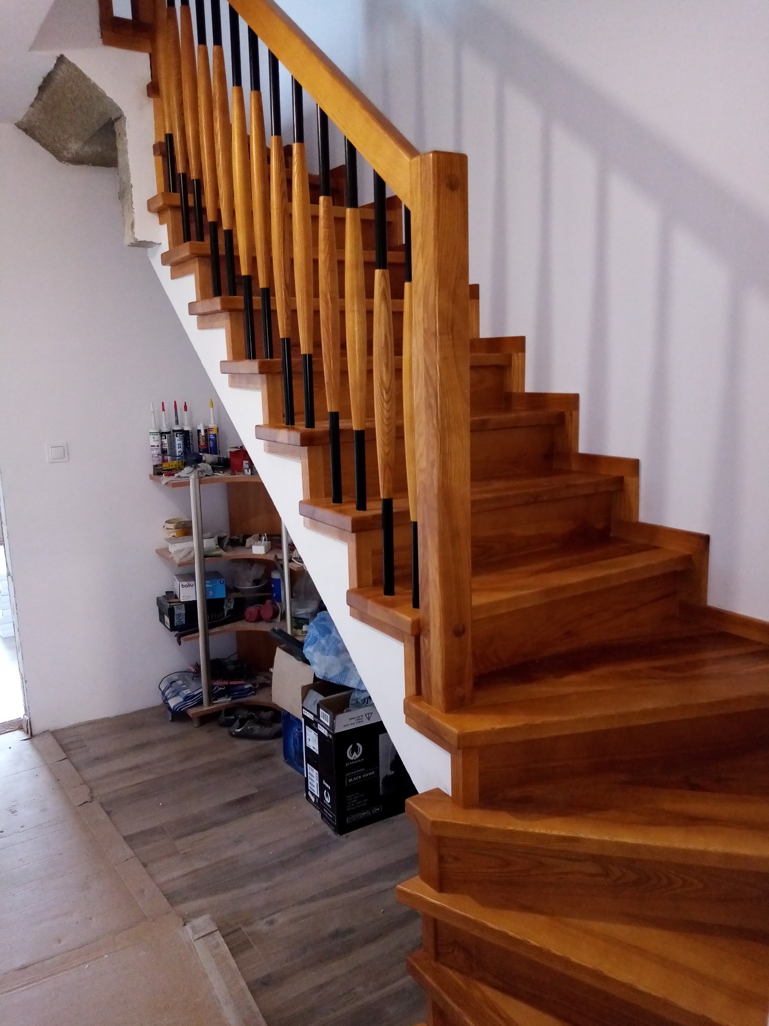 Nowoczesne schody z drewna z czarnymi, pionowymi elementami balustrady, widok na półki z narzędziami i materiałami budowlanymi w tle.