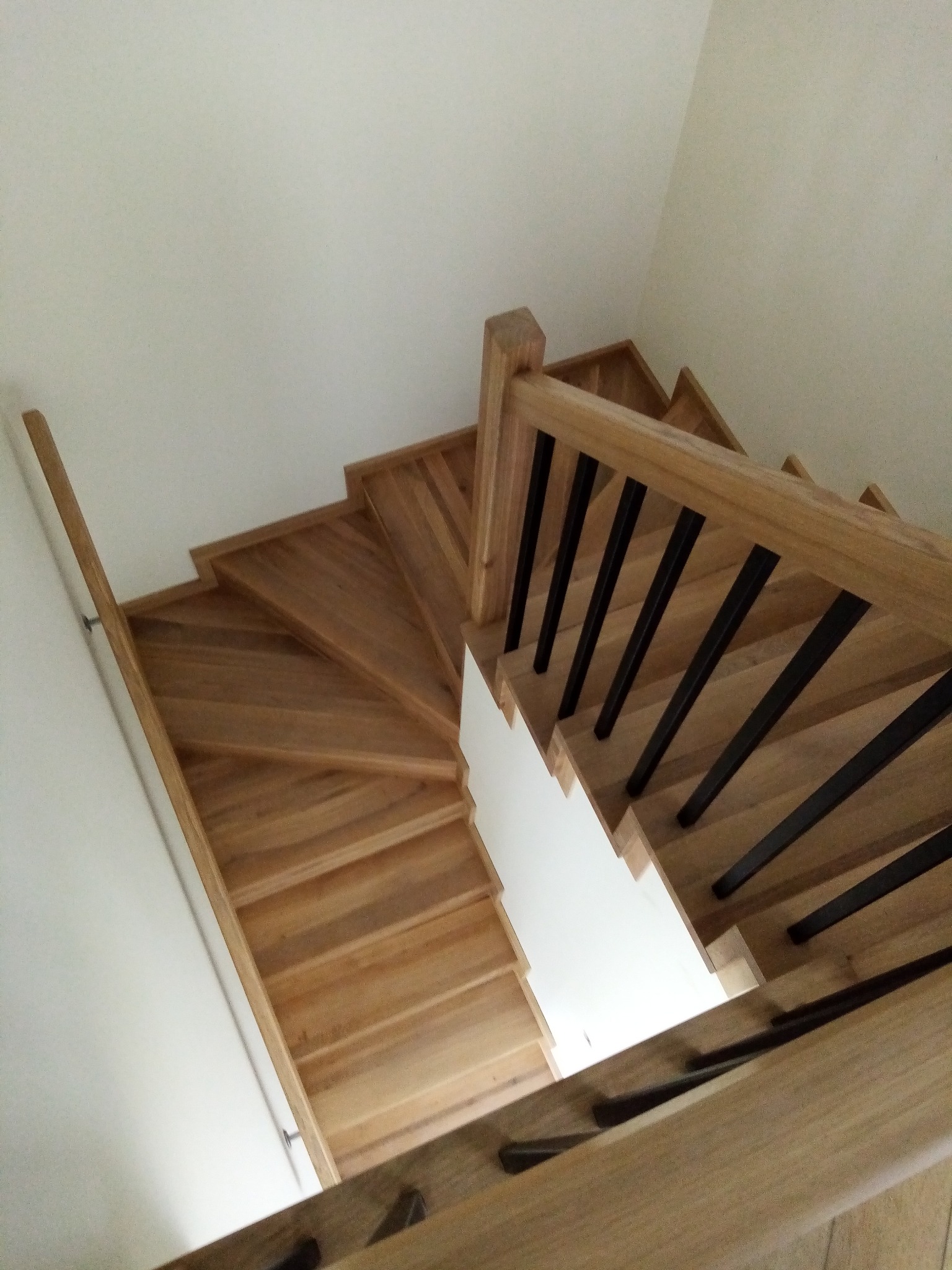 Drewniane schody z czarnymi metalowymi tralkami, widok z góry, skręcające w lewo, z jasnym oświetleniem w dolnej części biegu.