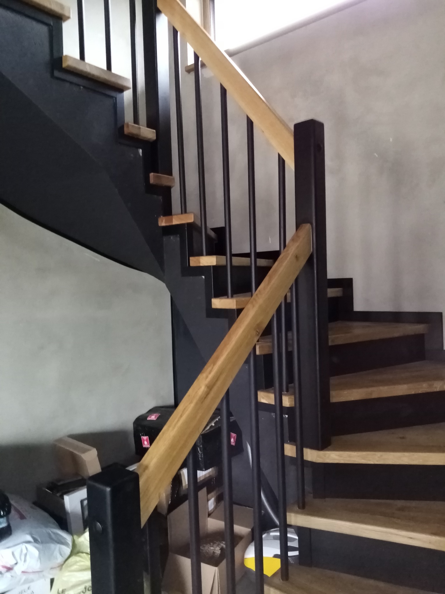 Nowoczesne schody z drewnianymi stopniami i czarną konstrukcją oraz jasną drewnianą poręczą, widoczne elementy montażowe i kartony w tle.