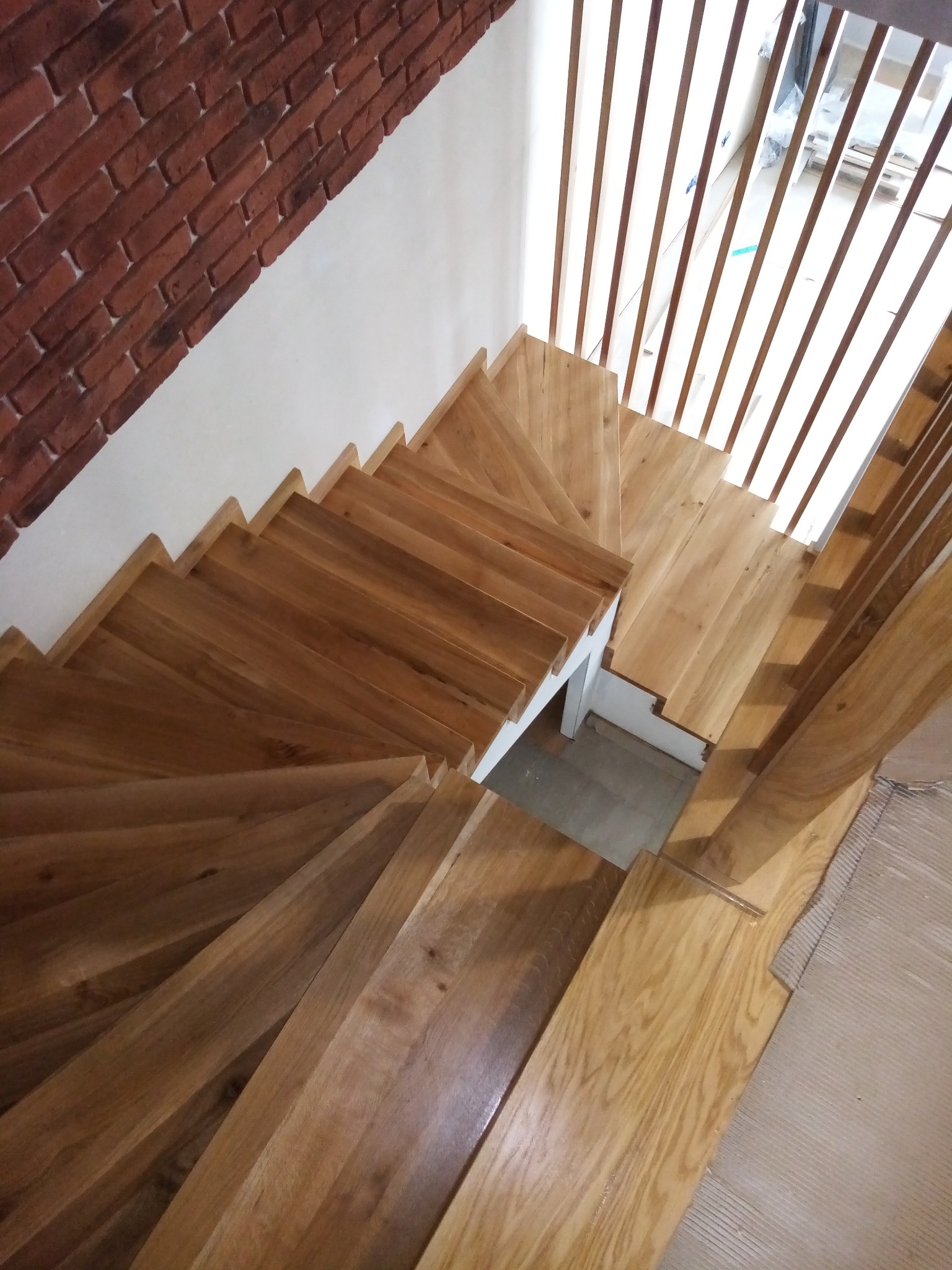 Widok z góry na drewniane schody z jasnego drewna, z minimalistyczną balustradą z pionowych listew i fragmentem ściany z cegły.