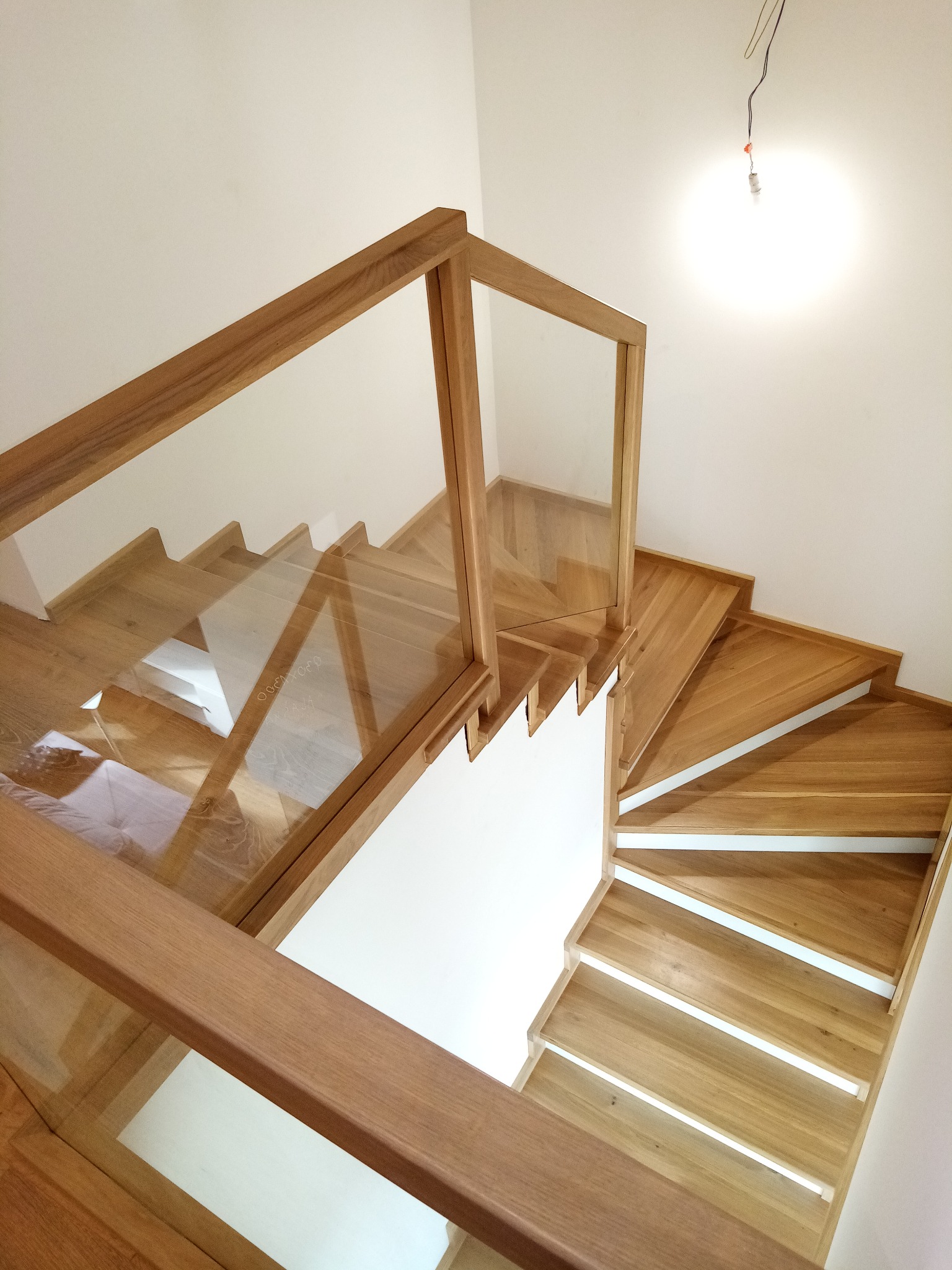 Nowoczesne schody z drewna i szkła, widok z góry, jasne oświetlenie, białe ściany, minimalistyczny design, widoczne przewody elektryczne w górnym rogu.