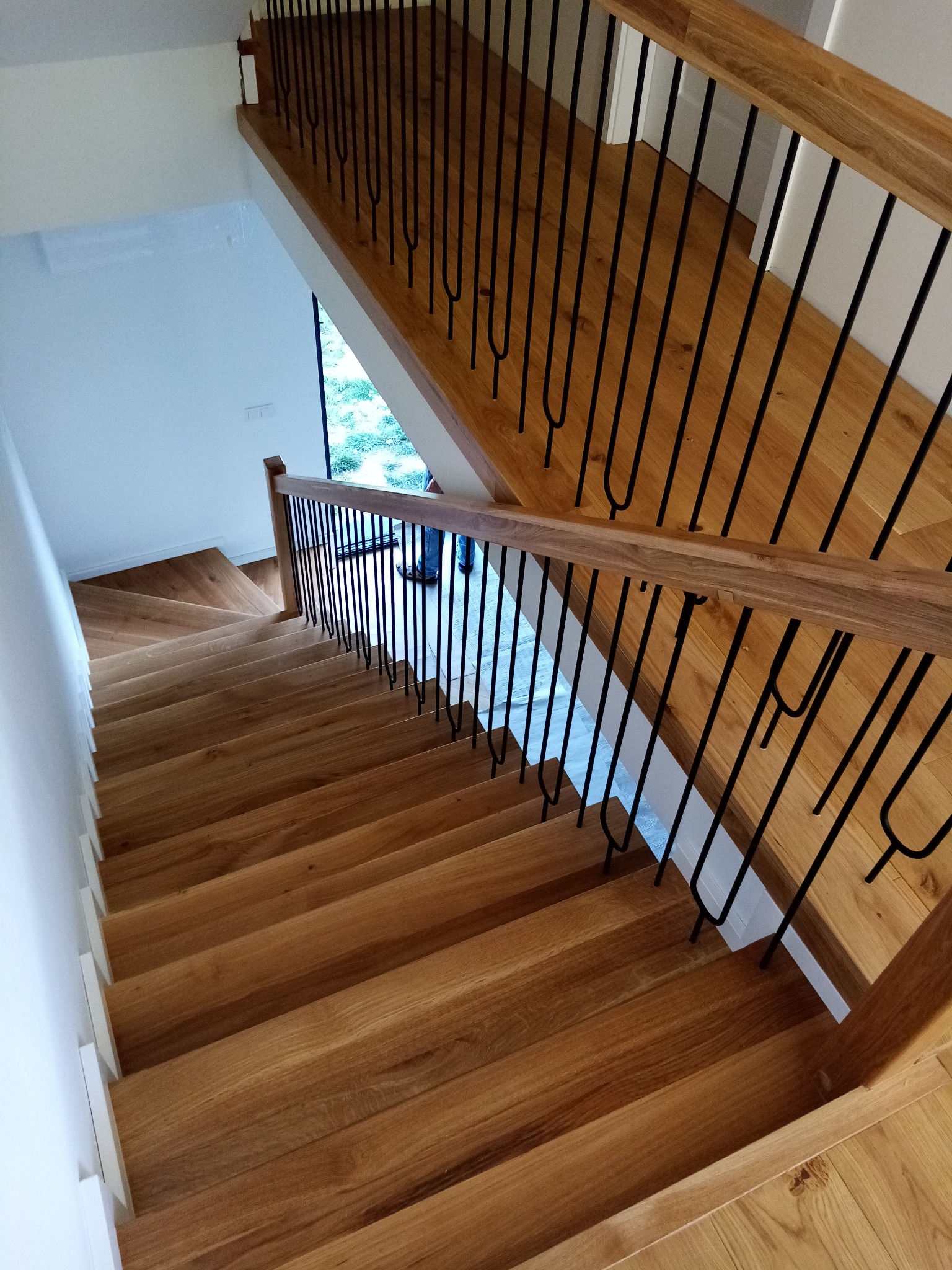 Widok z góry na jasne, drewniane stopnie z czarnymi, metalowymi szprosami w balustradzie i drewnianą poręczą, prowadzące w dół w nowoczesnym wnętrzu.