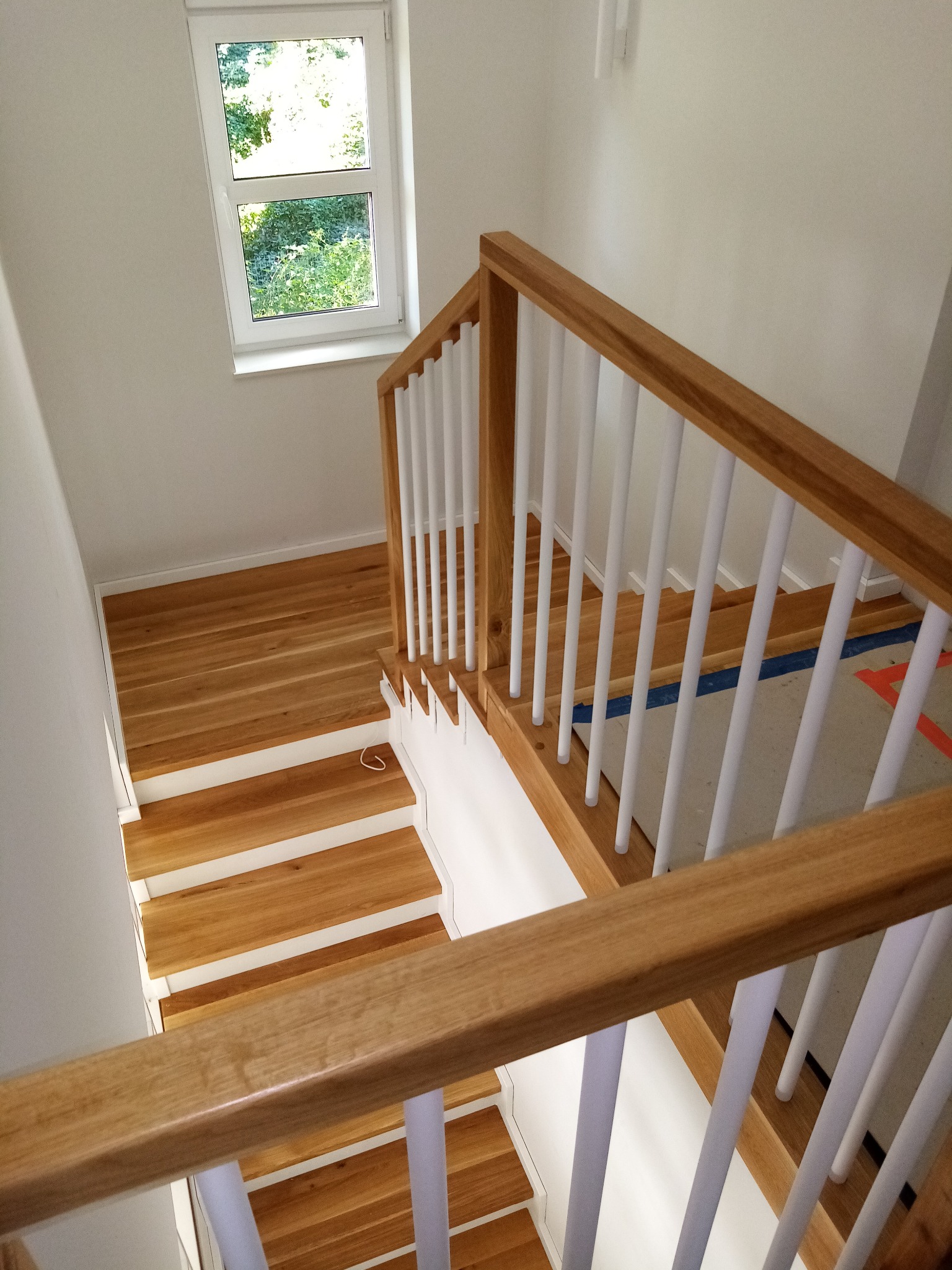 Widok z góry na jasne schody drewniane z białymi balustradami i drewnianą poręczą, częściowo zakryte folią ochronną na podłodze, okno w tle.