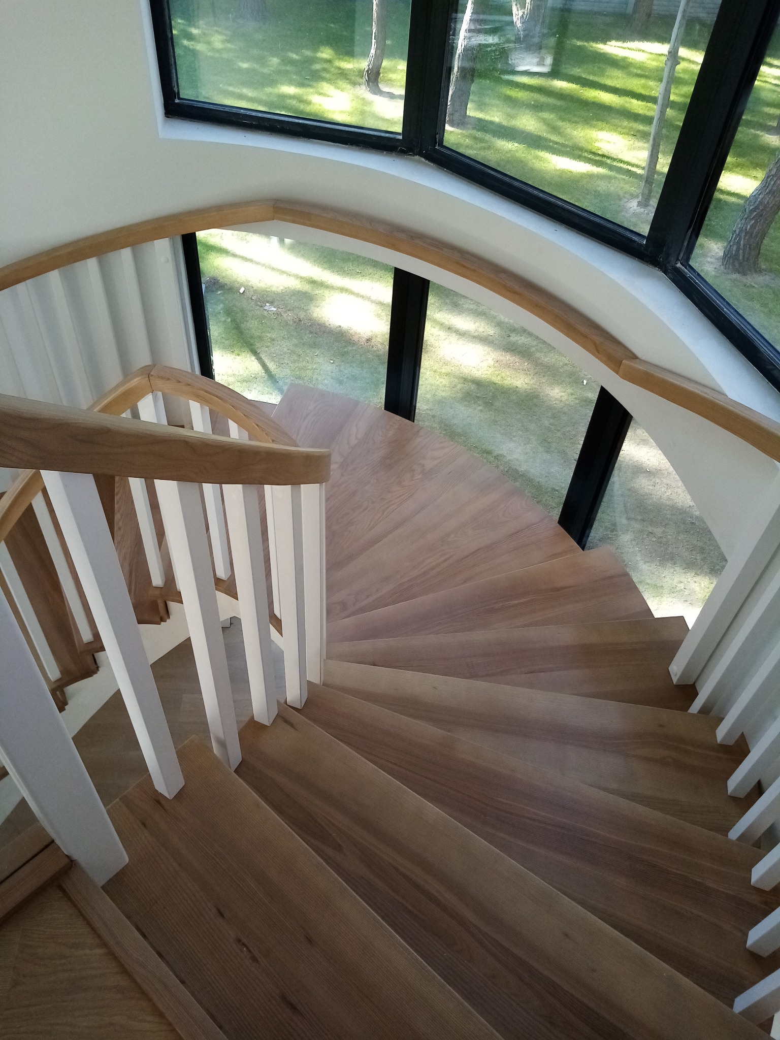 Spiralne schody z jasnego drewna z białymi balustradami, widok z góry na tle okna z widokiem na trawnik