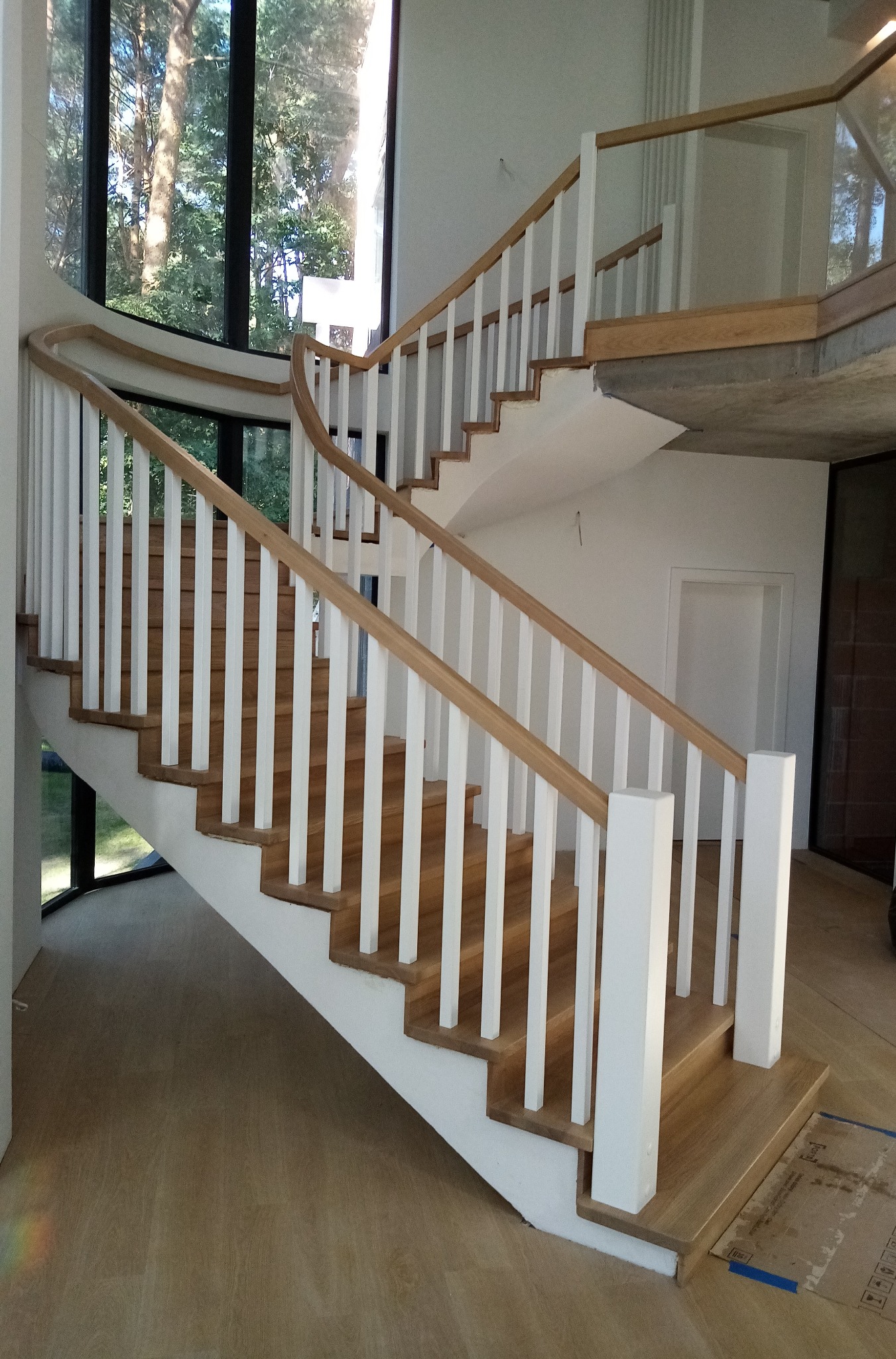 Nowoczesne, zakrzywione schody z drewnianymi stopniami i białymi balustradami w jasnym wnętrzu z dużym oknem.