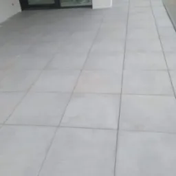 Jasnoszare, kwadratowe płytki ułożone na zewnątrz, widoczne przesuwne drzwi balkonowe i szklana balustrada.