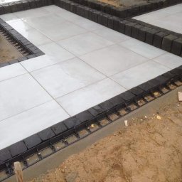 Z.U. Robert Mikulski - Układanie kostki brukowej - Białe płyty tarasowe obramowane ciemną kostką granitową, widoczne mocowania i przygotowane podłoże, etap prac wykończeniowych.