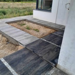 Z.U. Robert Mikulski - Układanie kostki brukowej - Układanie ciemnej kostki brukowej z granitowymi obrzeżami przy ścianie domu, z wydzielonym miejscem na rabatę kwiatową wypełnioną piaskiem i młodymi roślinami.