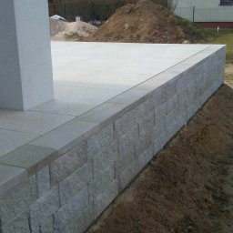 Z.U. Robert Mikulski - Układanie kostki brukowej - Wykończony taras z jasnoszarej kostki, obrzeża i ściana oporowa z szarego kamienia łupanego, widok na posesję z domem w tle.