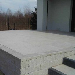 Z.U. Robert Mikulski - Układanie kostki brukowej - Jasnoszara kostka granitowa tworząca taras z niskim murkiem i schodami, widok z dołu na tle nowoczesnego domu.