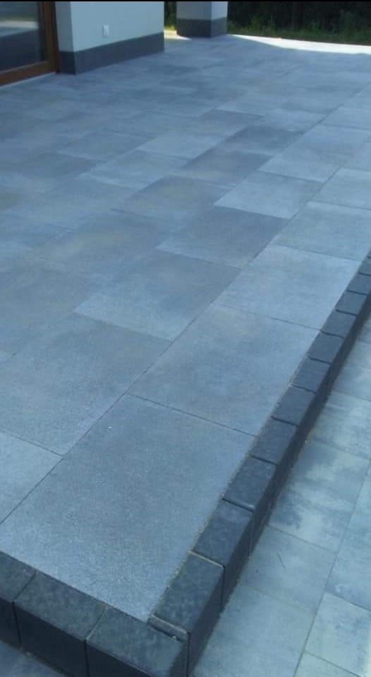 Szary, prostokątny taras z kostki brukowej, obrzeża wykończone ciemniejszymi kostkami, widok na budynek w tle.