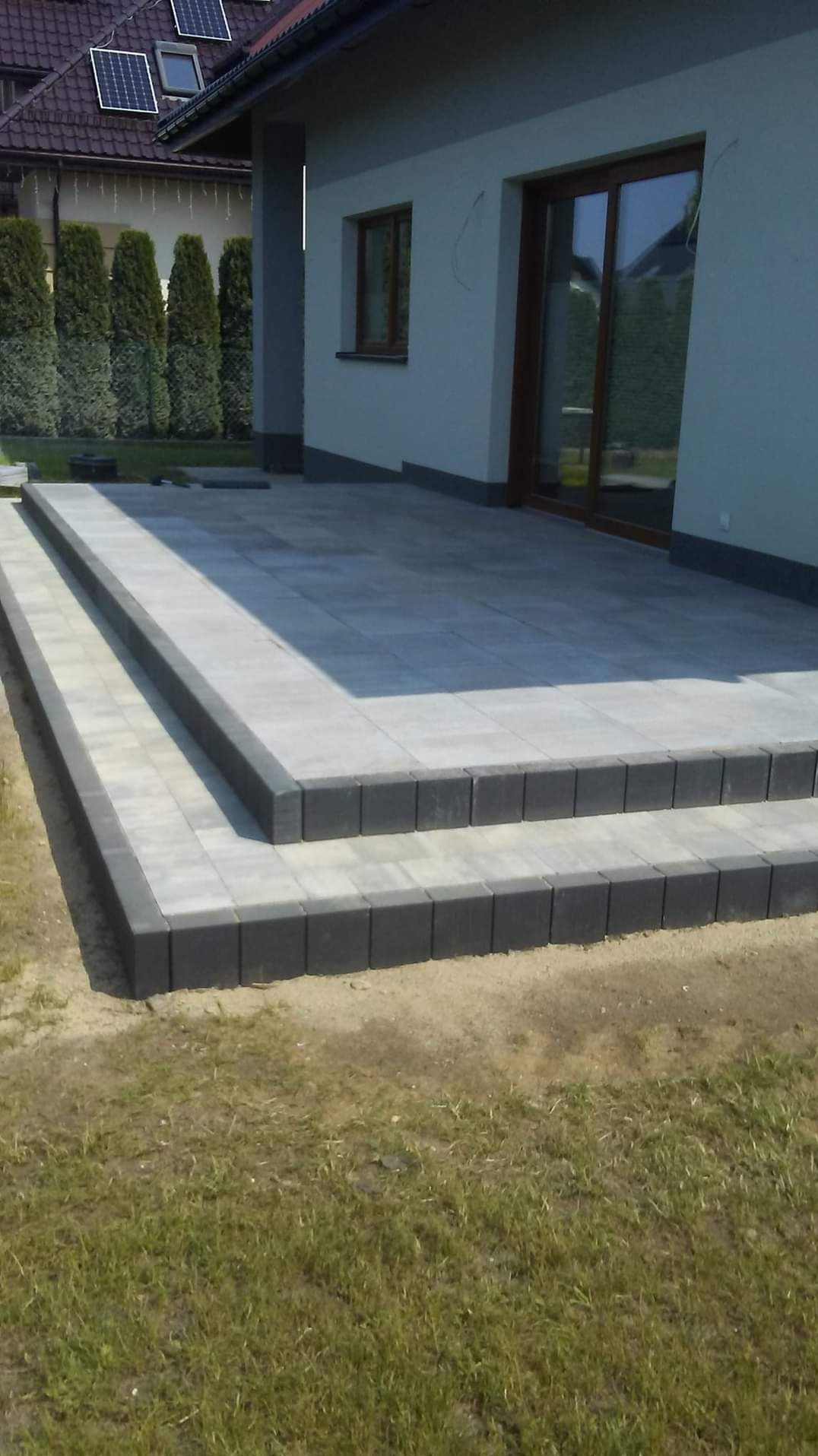 Nowoczesny taras z szarej kostki brukowej z dwoma stopniami, obrzeża z ciemnej kostki, trawnik z przodu, dom z szarym tynkiem w tle.