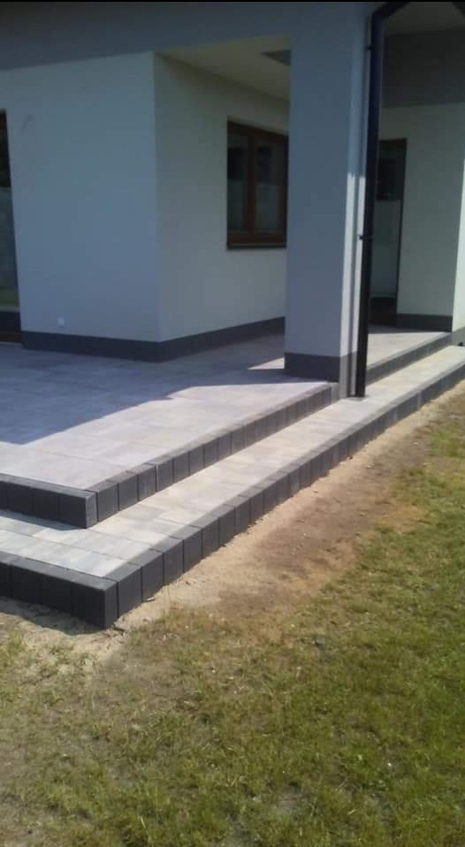 Schody i taras wyłożone szarą kostką brukową z ciemnymi obrzeżami, widok z poziomu trawnika, w tle fragment elewacji budynku.