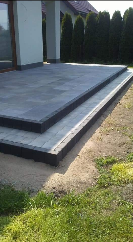 Nowoczesny taras z szarej kostki brukowej z czarnym obrzeżem, prowadzący do domu, otoczony trawnikiem i tujami.