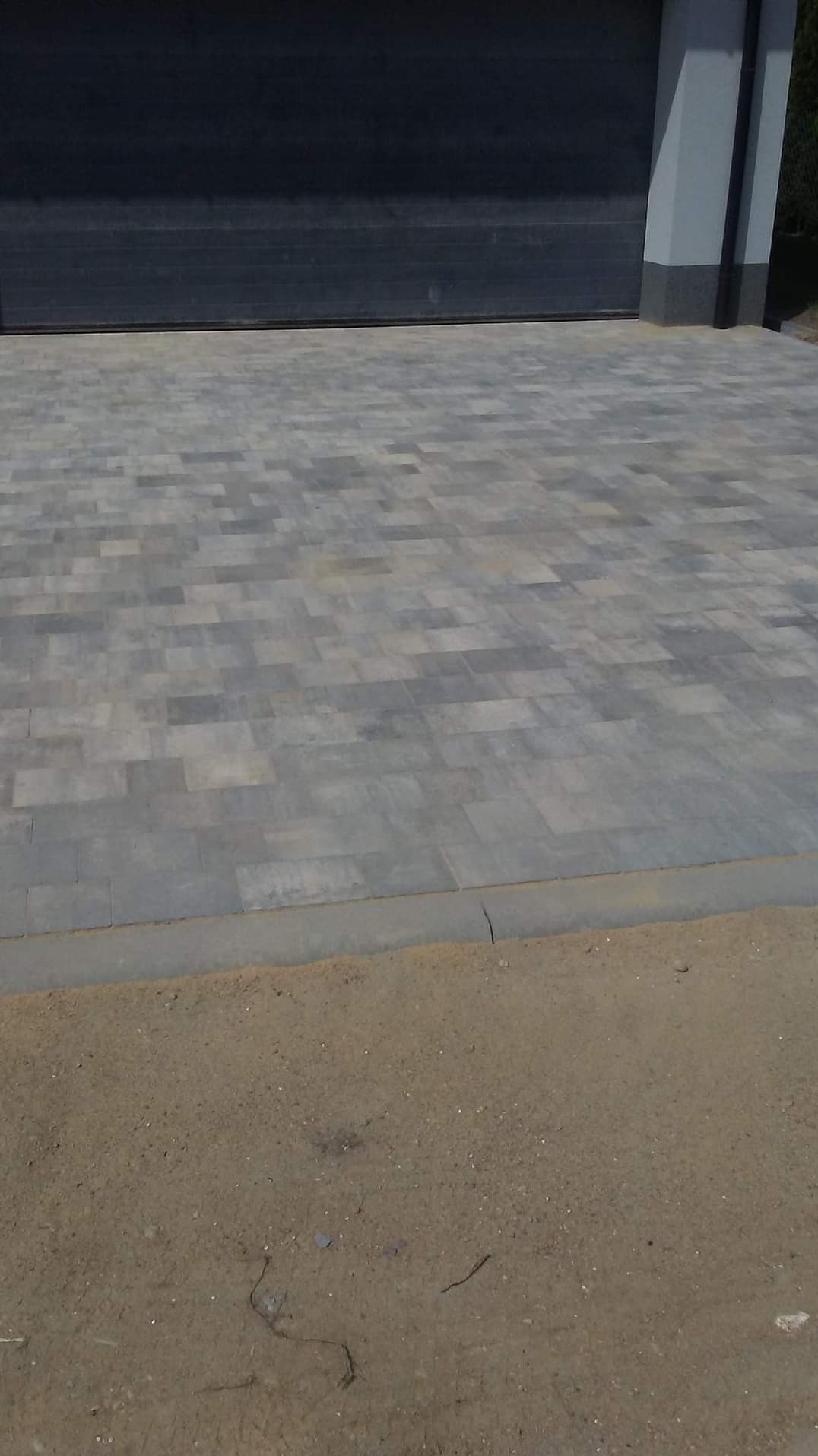 Ułożona szara kostka brukowa na podjeździe przed garażem z automatyczną bramą, widoczne obrzeża i fragment nieutwardzonego terenu.