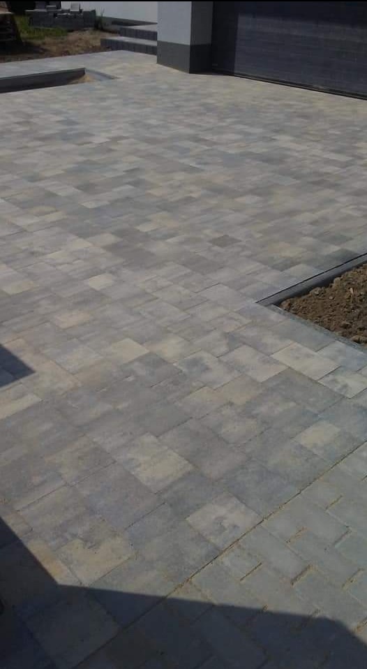 Szara kostka brukowa ułożona na podjeździe, widoczne schody i brama garażowa w tle, świeżo wykończone obrzeże z ziemią.
