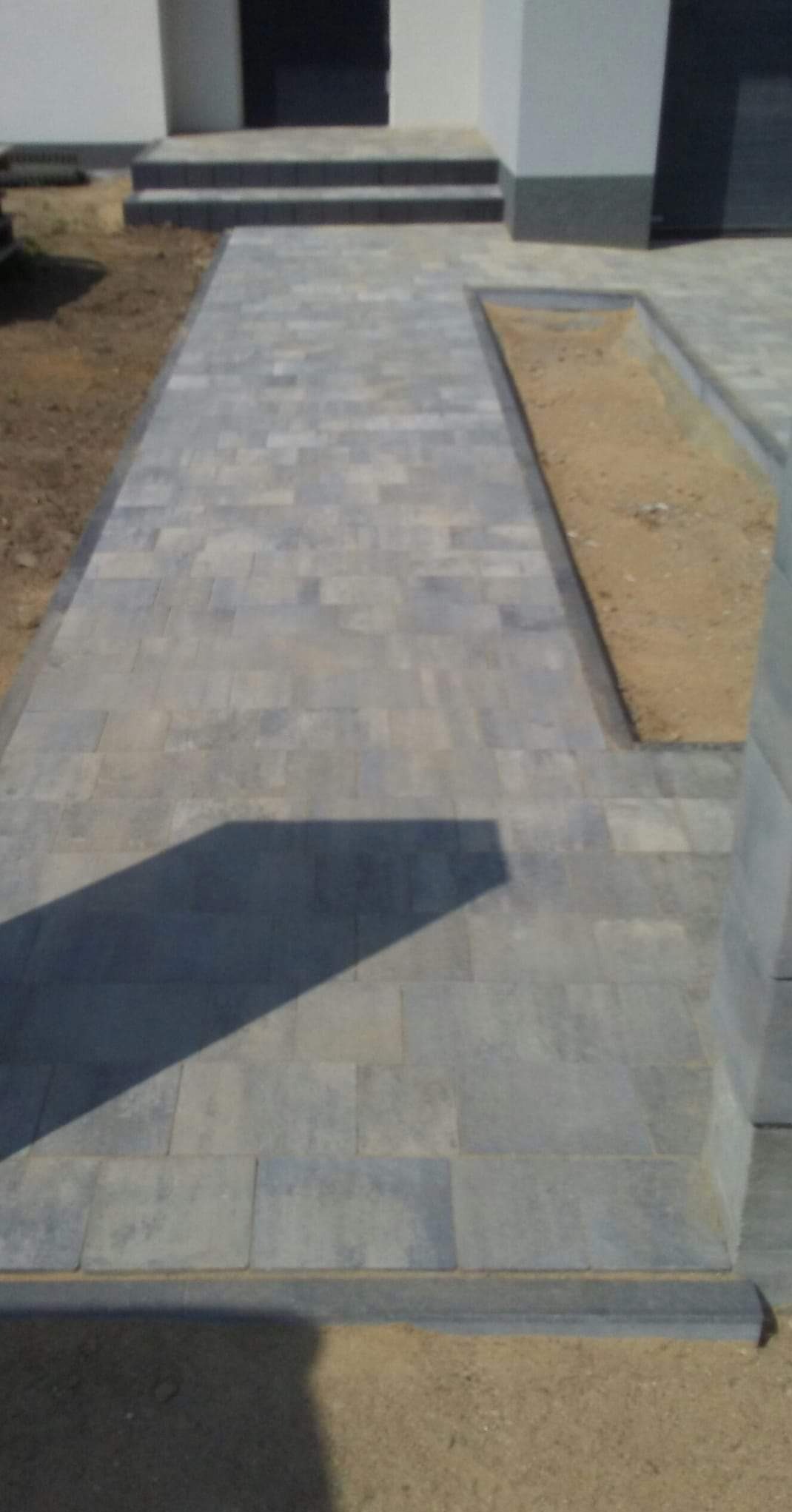 Ułożona szara kostka brukowa prowadząca do wejścia budynku z widocznym fragmentem rabaty ziemnej obok ścieżki.