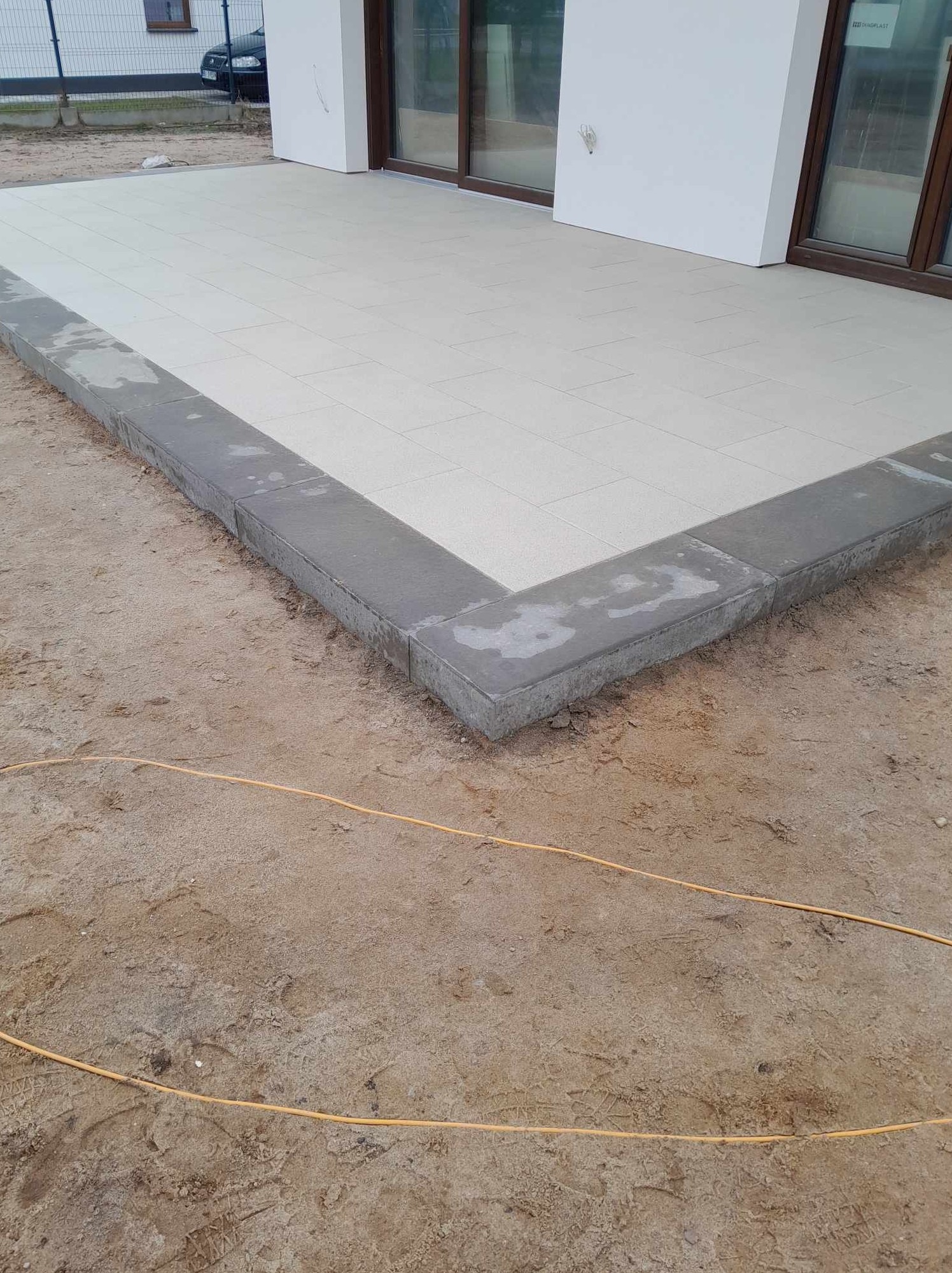 Jasne płyty tarasowe obramowane szarym betonowym obrzeżem, widoczne ślady wilgoci na obrzeżu, piaszczyste podłoże z żółtym przewodem, fragment budynku z brązowymi oknami.