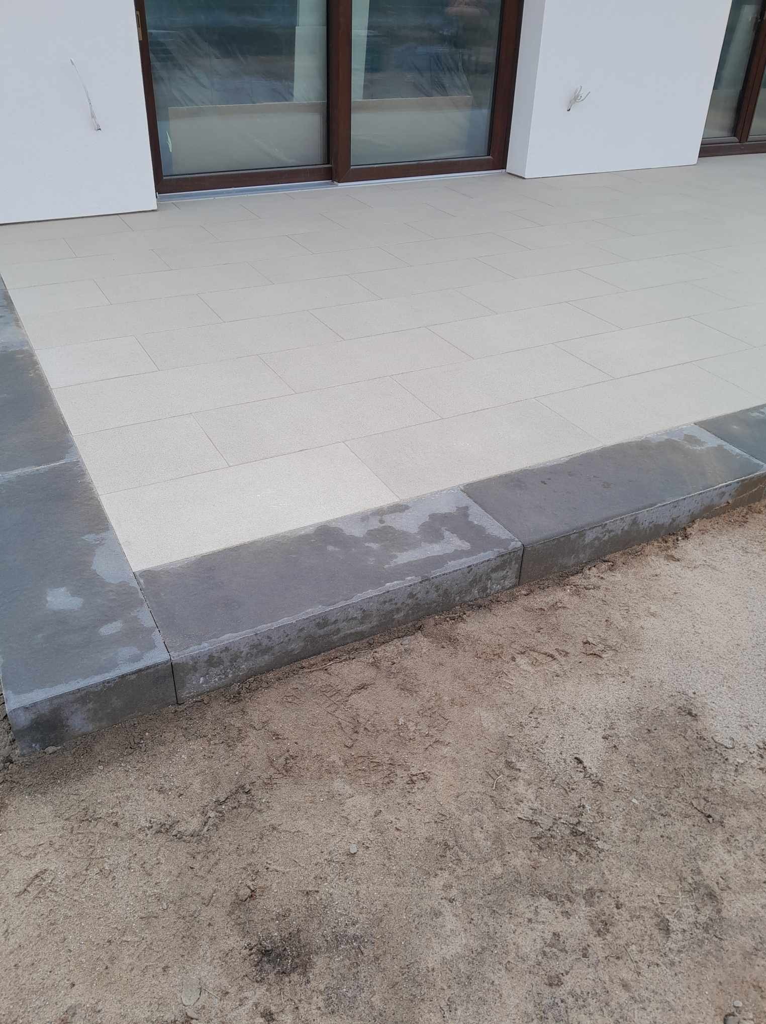 Wykończony taras z jasnych, prostokątnych płyt, obrzeża z ciemnoszarego kamienia, widoczne ślady wilgoci na obrzeżach, surowy grunt w tle.