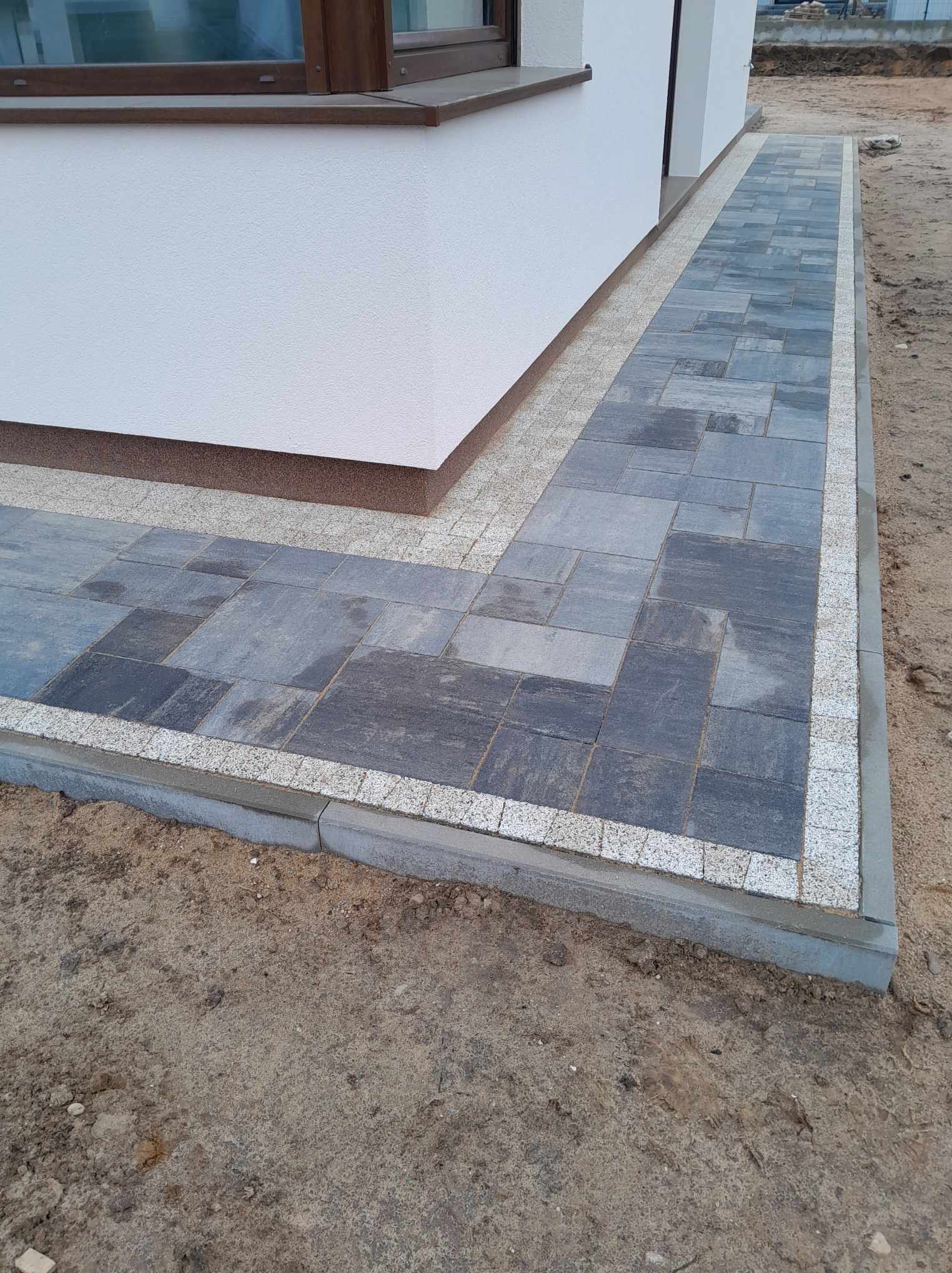 Świeżo ułożony chodnik z szarej kostki brukowej obramowany jasną kostką granitową, przylegający do białej ściany budynku z brązowymi elementami wykończeniowymi