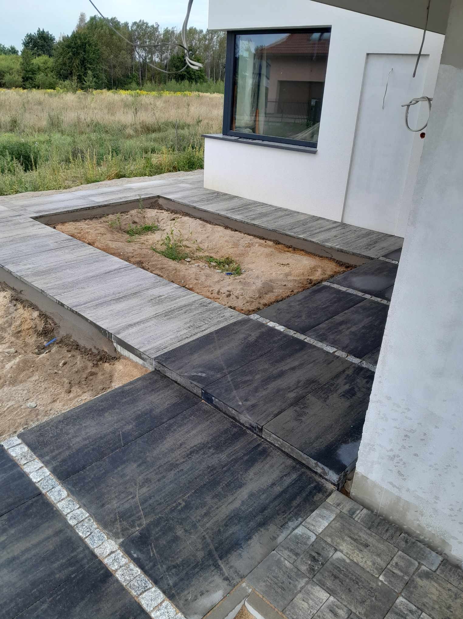 Układanie ciemnej kostki brukowej z granitowymi obrzeżami przy ścianie domu, z wydzielonym miejscem na rabatę kwiatową wypełnioną piaskiem i młodymi roślinami.