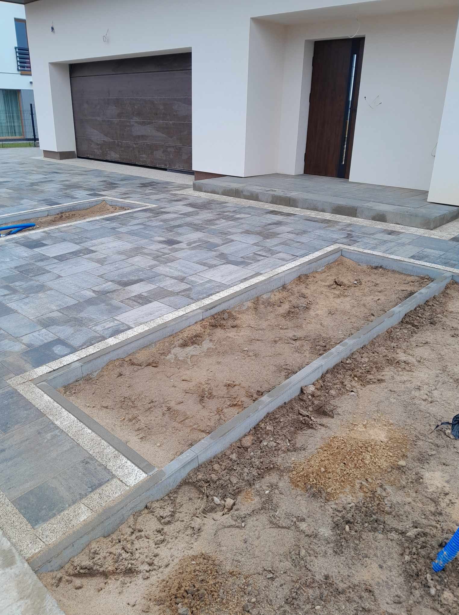 Przygotowanie podłoża pod rabatę kwiatową obok podjazdu z szarej kostki brukowej, widoczne obrzeża i piasek wewnątrz.