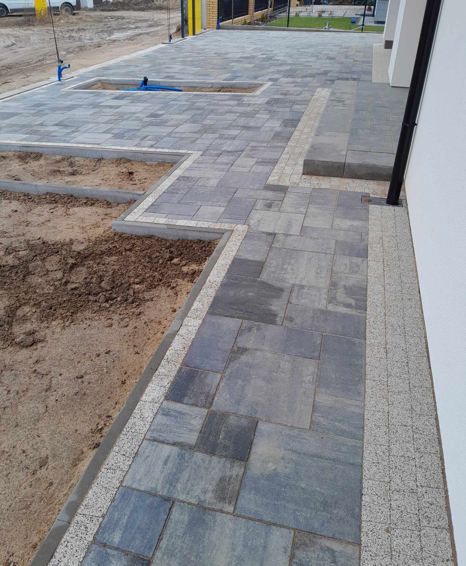 Szara kostka brukowa ułożona w geometryczny wzór, obrzeża z jasnej kostki, fragmenty terenu przygotowane pod dalsze prace, widoczny fragment budynku i rury spustowej.