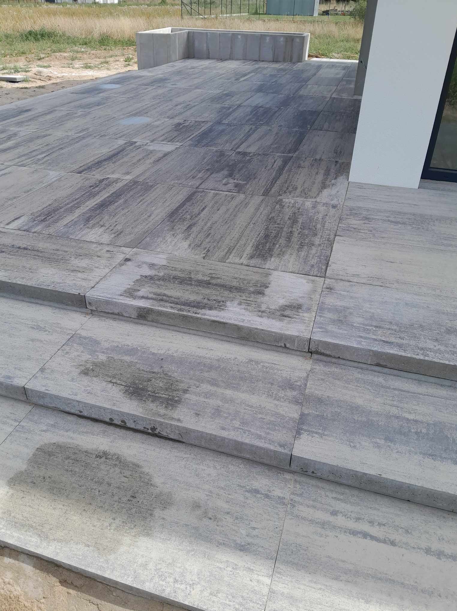 Nowoczesny taras z szarych, prostokątnych płyt z subtelnym wzorem, prowadzący do budynku z białym filarem i dużym oknem, z widocznymi schodami i elementami krajobrazu w tle.