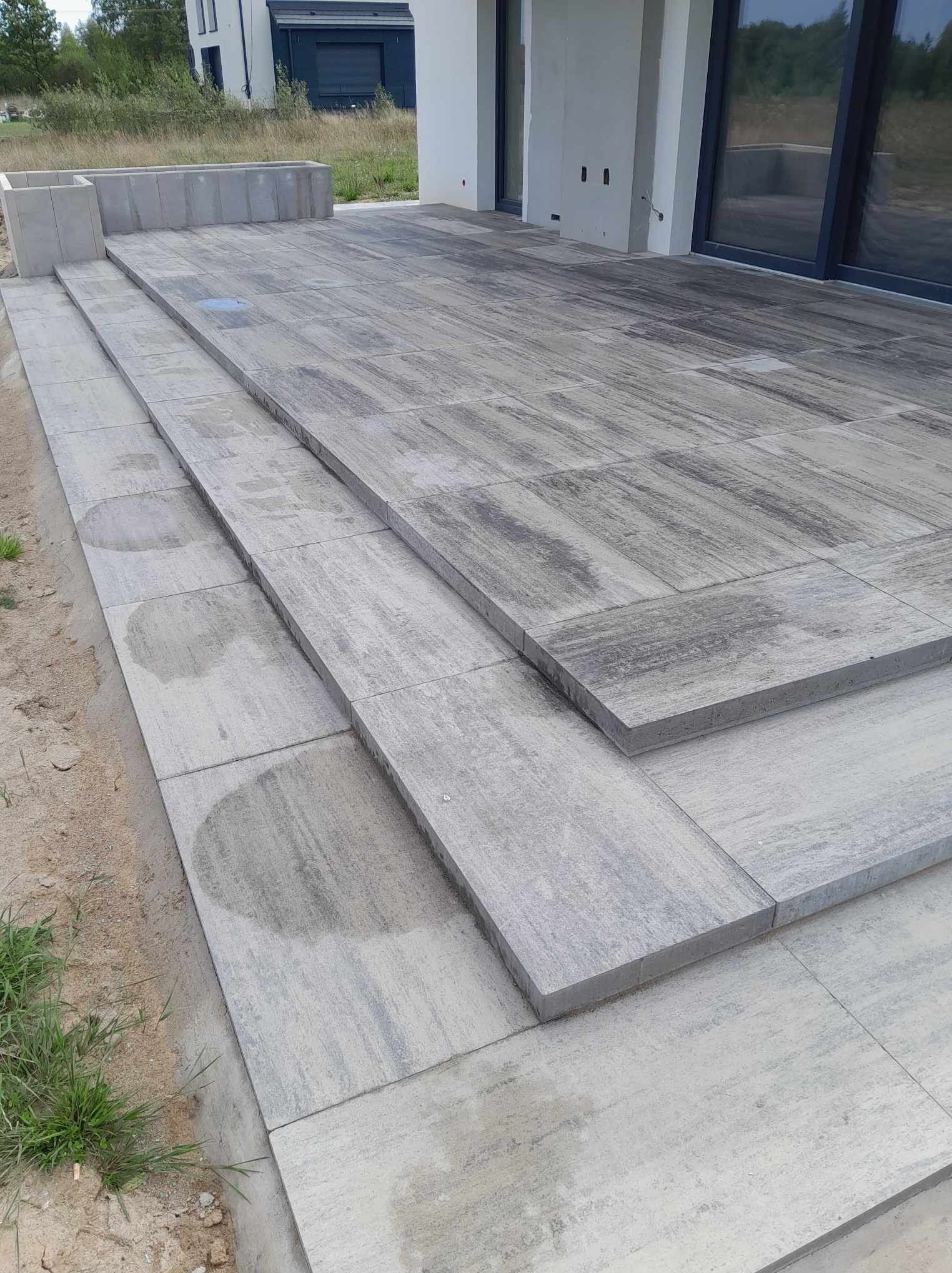 Schody i taras wyłożone szarą kostką o nieregularnym wzorze, z widocznymi śladami wilgoci, przylegające do nowoczesnego domu z dużymi oknami.
