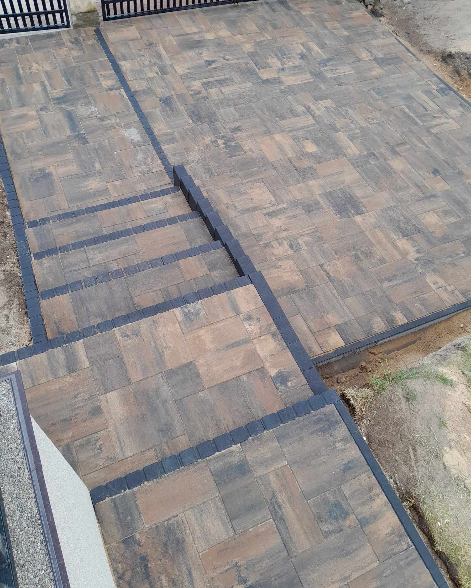 Ułożona nawierzchnia z kostki brukowej w odcieniach brązu i szarości, z obrzeżami z ciemnej kostki, prowadząca do bramy i schodów.