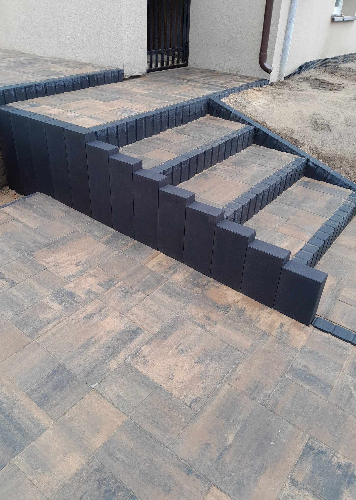 Schody zewnętrzne wyłożone kostką brukową w odcieniach szarości i beżu, obrzeża z ciemnoszarej kostki, widok z góry.