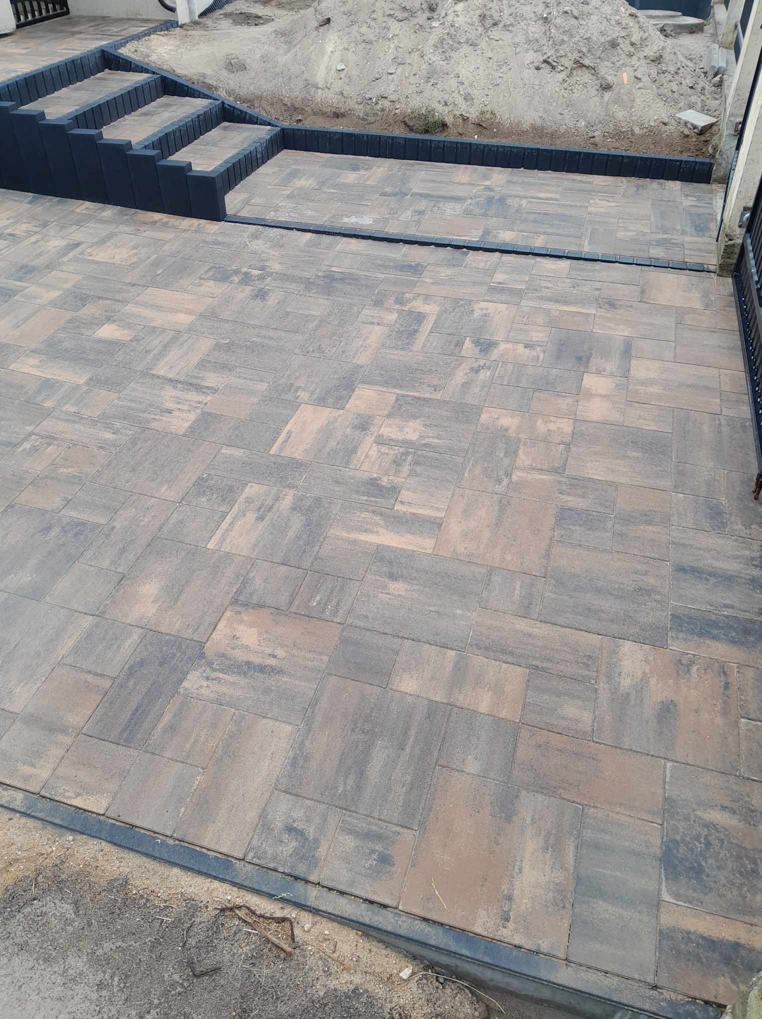 Ułożona kostka brukowa w odcieniach brązu i szarości z czarnymi obrzeżami i schodami, widoczny nasyp ziemi w tle.