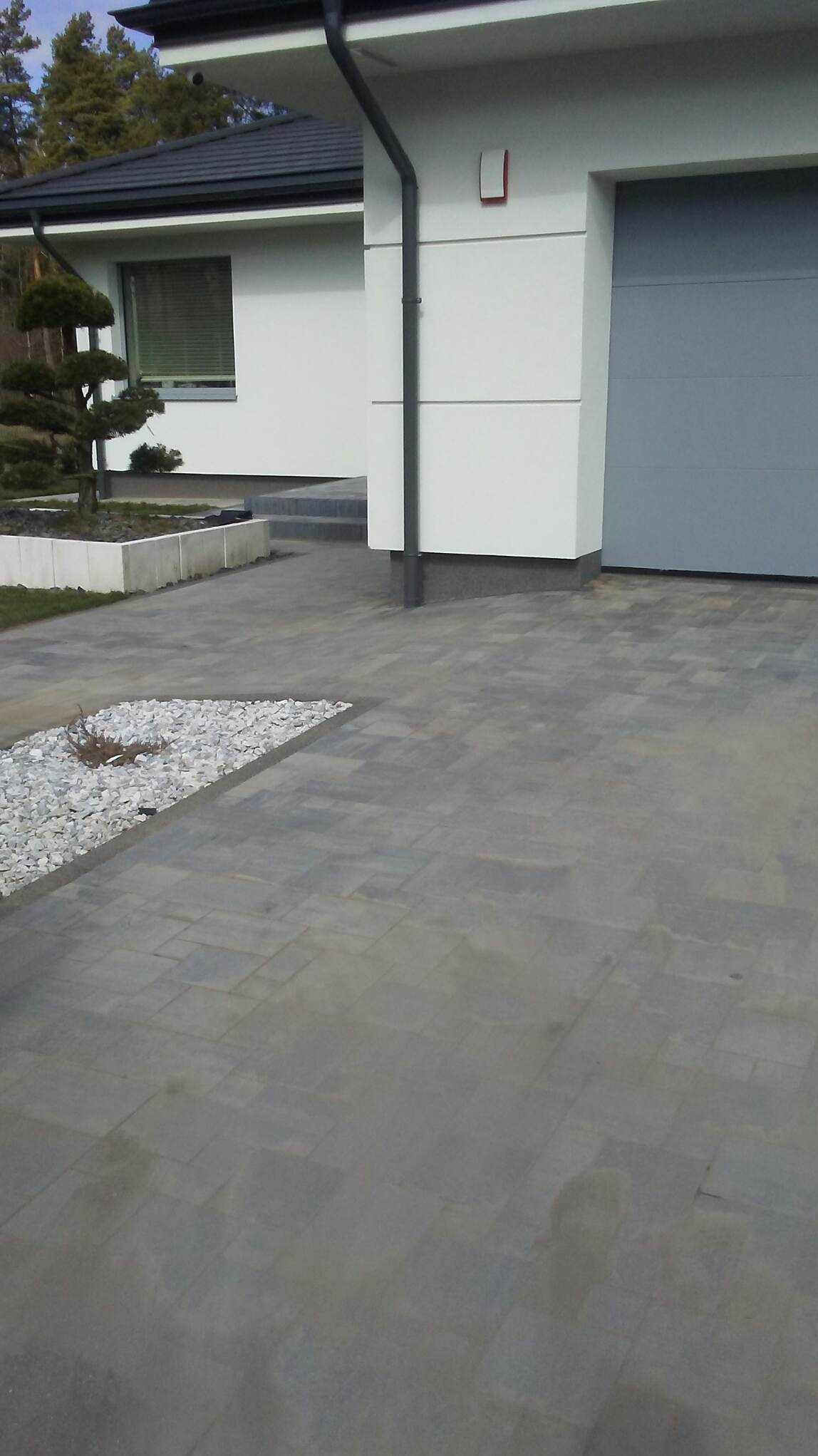 Szara nawierzchnia z kostki brukowej przed nowoczesnym domem z garażem, ozdobny klomb z białymi kamieniami i małym drzewkiem bonsai.