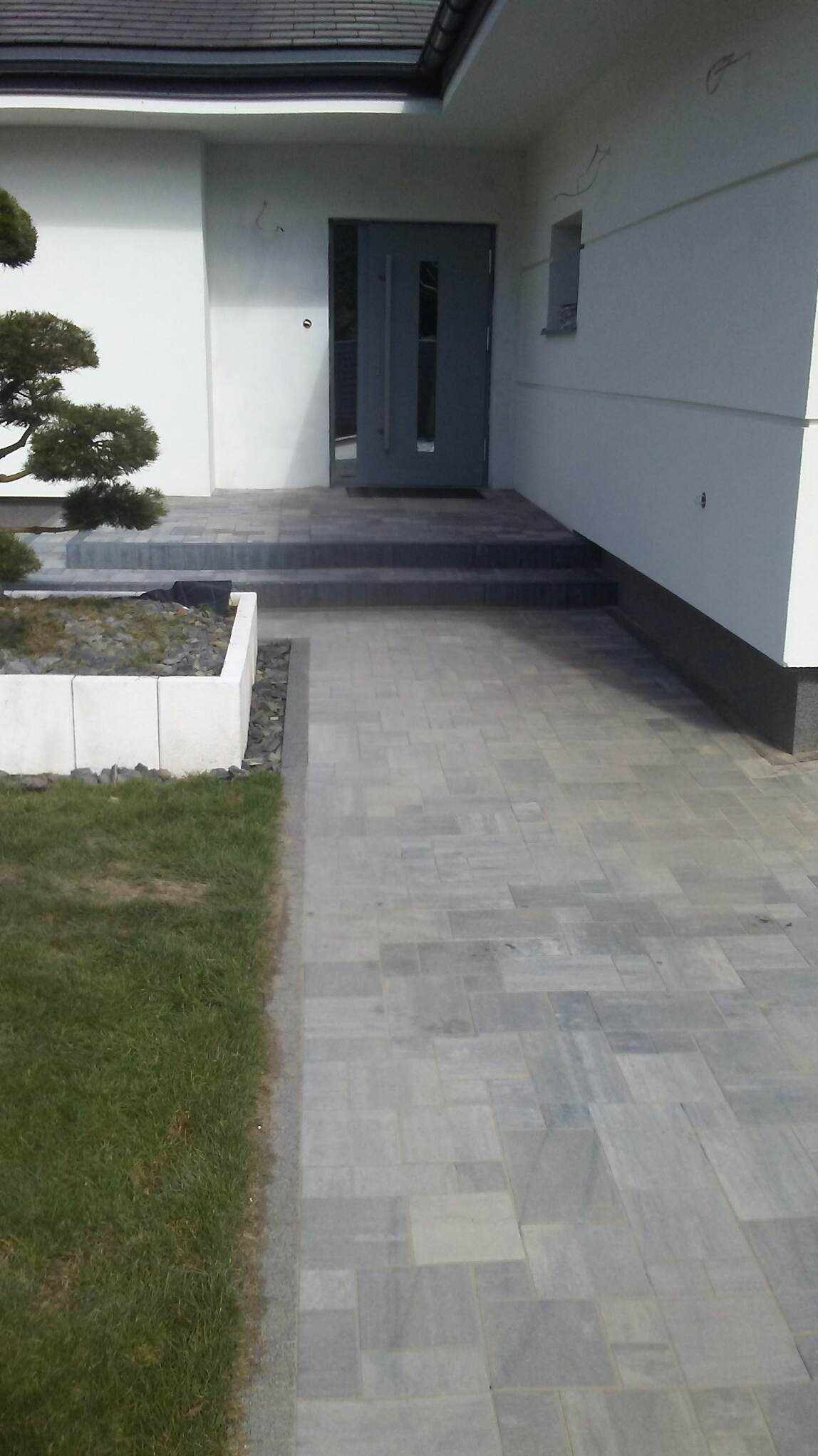 Szara kostka brukowa prowadząca do wejścia domu z szarymi drzwiami, obok trawnik i rabata z kamieniami.