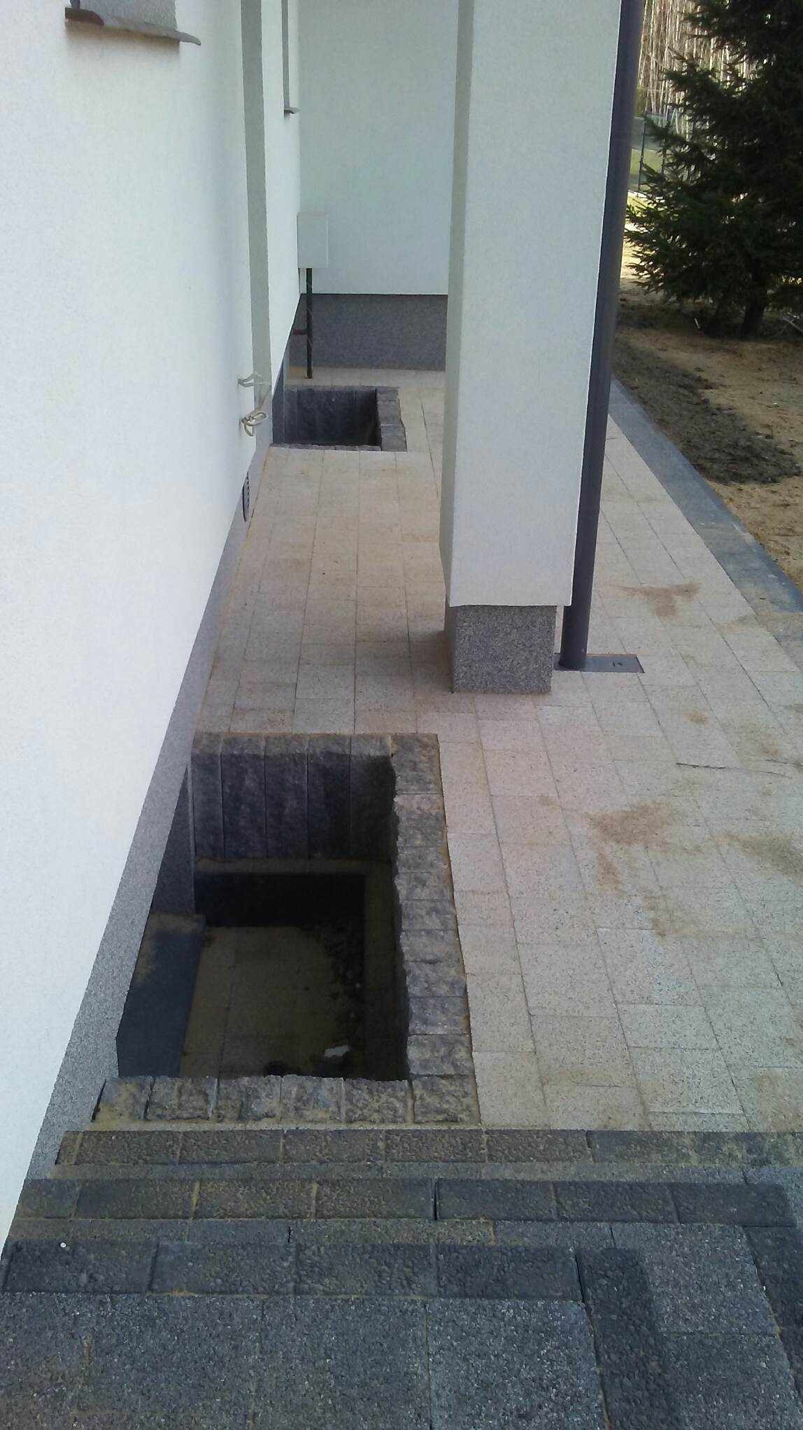 Wykończona ścieżka z jasnoszarej kostki brukowej prowadząca wzdłuż białej ściany budynku, z ciemnoszarymi obrzeżami i zagłębieniami wykończonymi ciemniejszą kostką, prawdopodobnie przeznaczonymi...