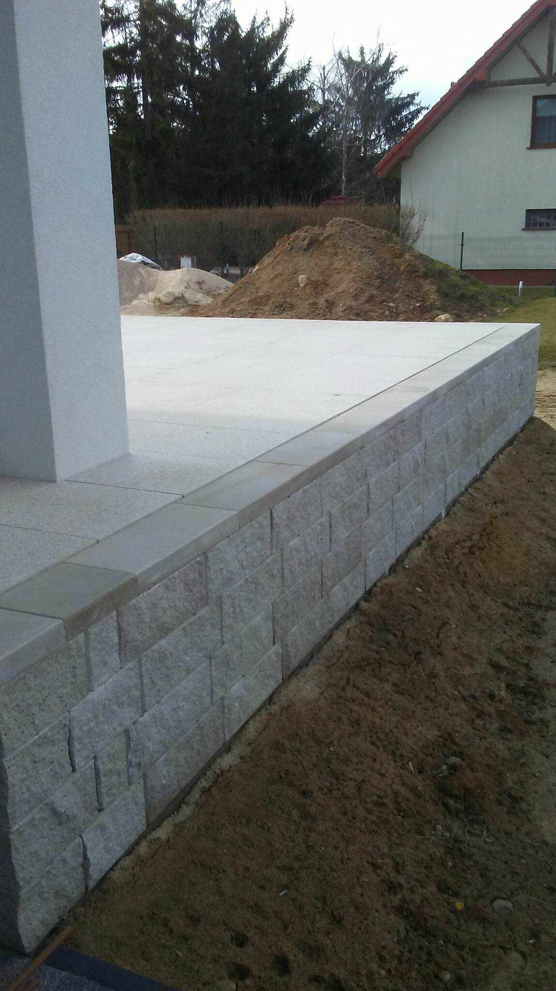 Wykończony taras z jasnoszarej kostki, obrzeża i ściana oporowa z szarego kamienia łupanego, widok na posesję z domem w tle.