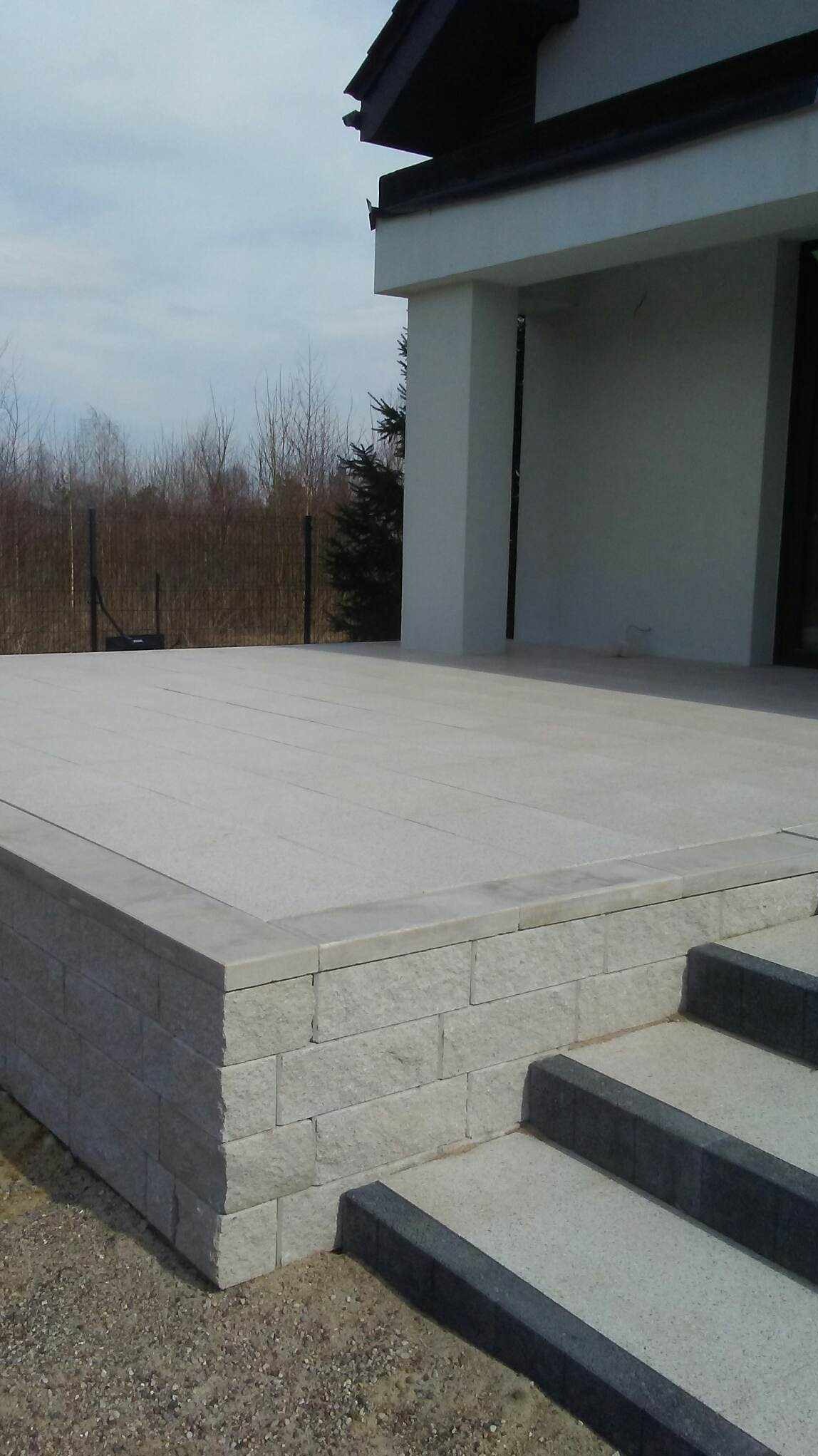 Jasnoszara kostka granitowa tworząca taras z niskim murkiem i schodami, widok z dołu na tle nowoczesnego domu.