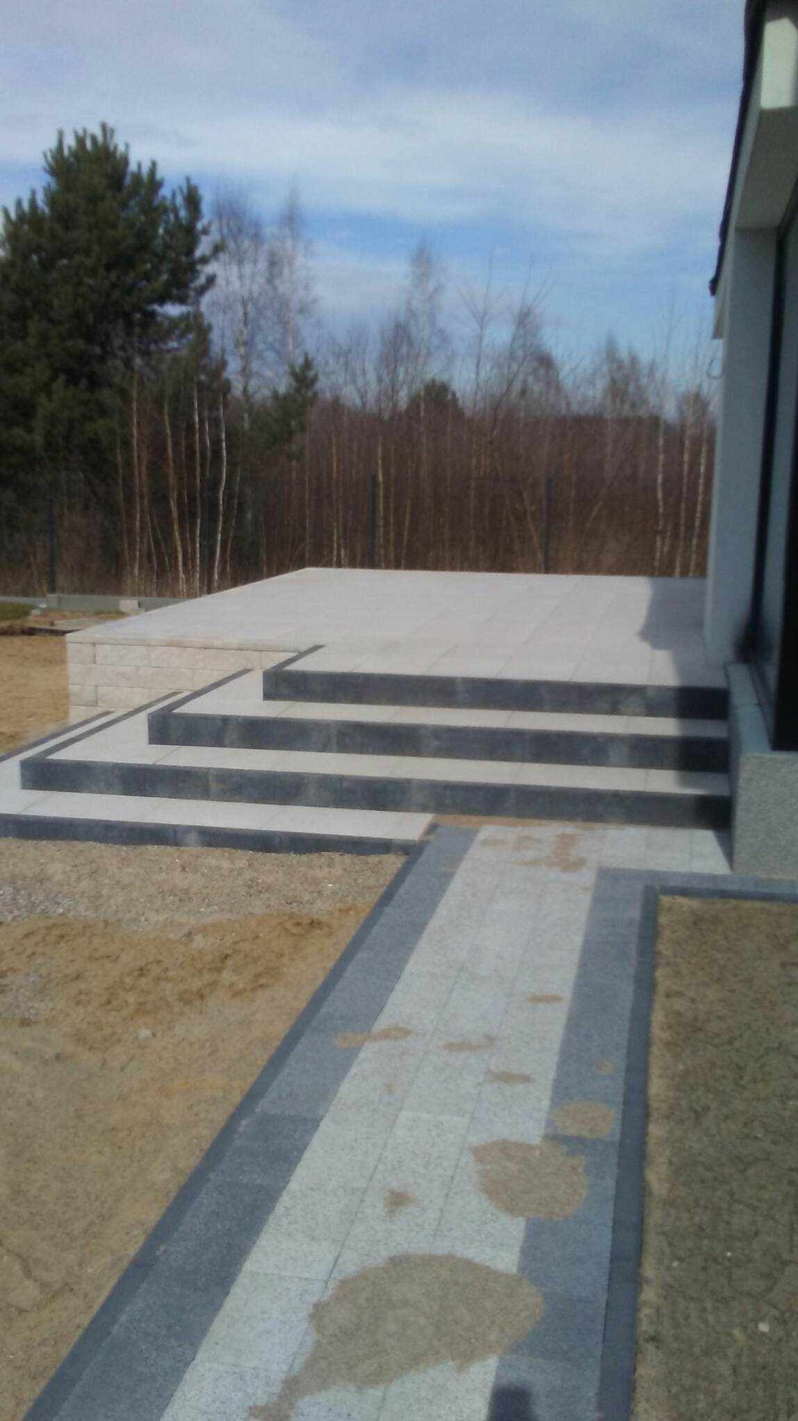 Schody i chodnik z jasnoszarej kostki z ciemnoszarym obramowaniem prowadzące do nowoczesnego domu, widok z perspektywy gruntu, w tle drzewa bez liści.