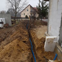 Usługi Elektryczne - Wykonany wykop pod niebieski przewód elektryczny na posesji w trakcie budowy, widoczny dom w tle i pochmurne niebo.