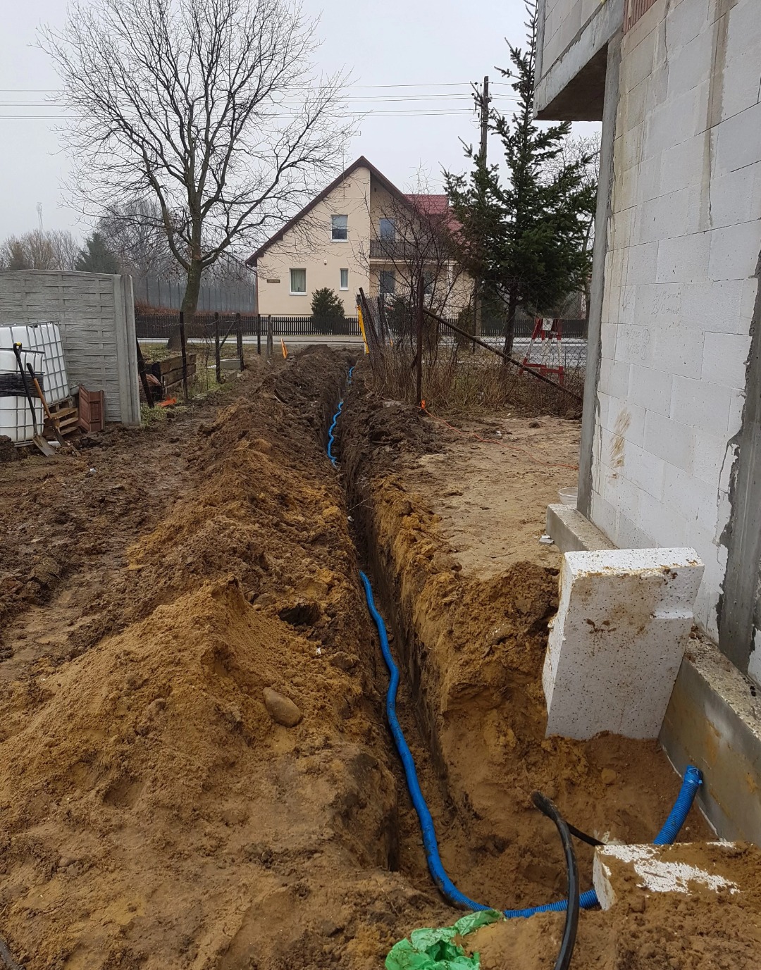 Wykonany wykop pod niebieski przewód elektryczny na posesji w trakcie budowy, widoczny dom w tle i pochmurne niebo.