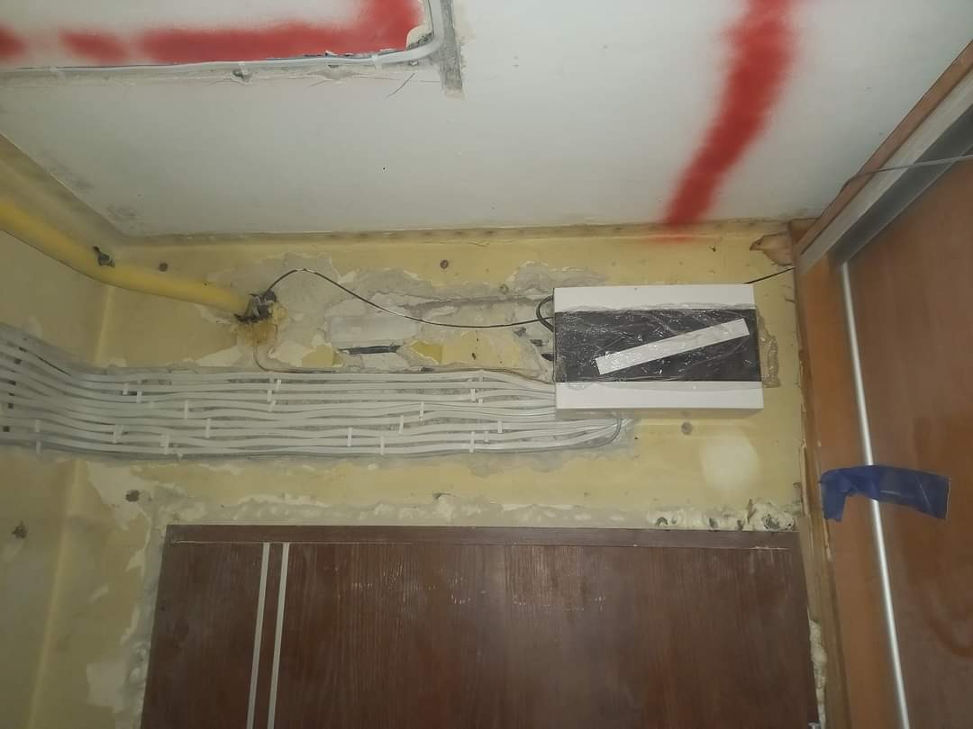 Ściana w trakcie remontu z widoczną puszką elektryczną zabezpieczoną folią i taśmą, oraz wiązką kabli w białych peszlach, prowadzących wzdłuż ściany. Widoczne ślady po kuciu w tynku i fragment...