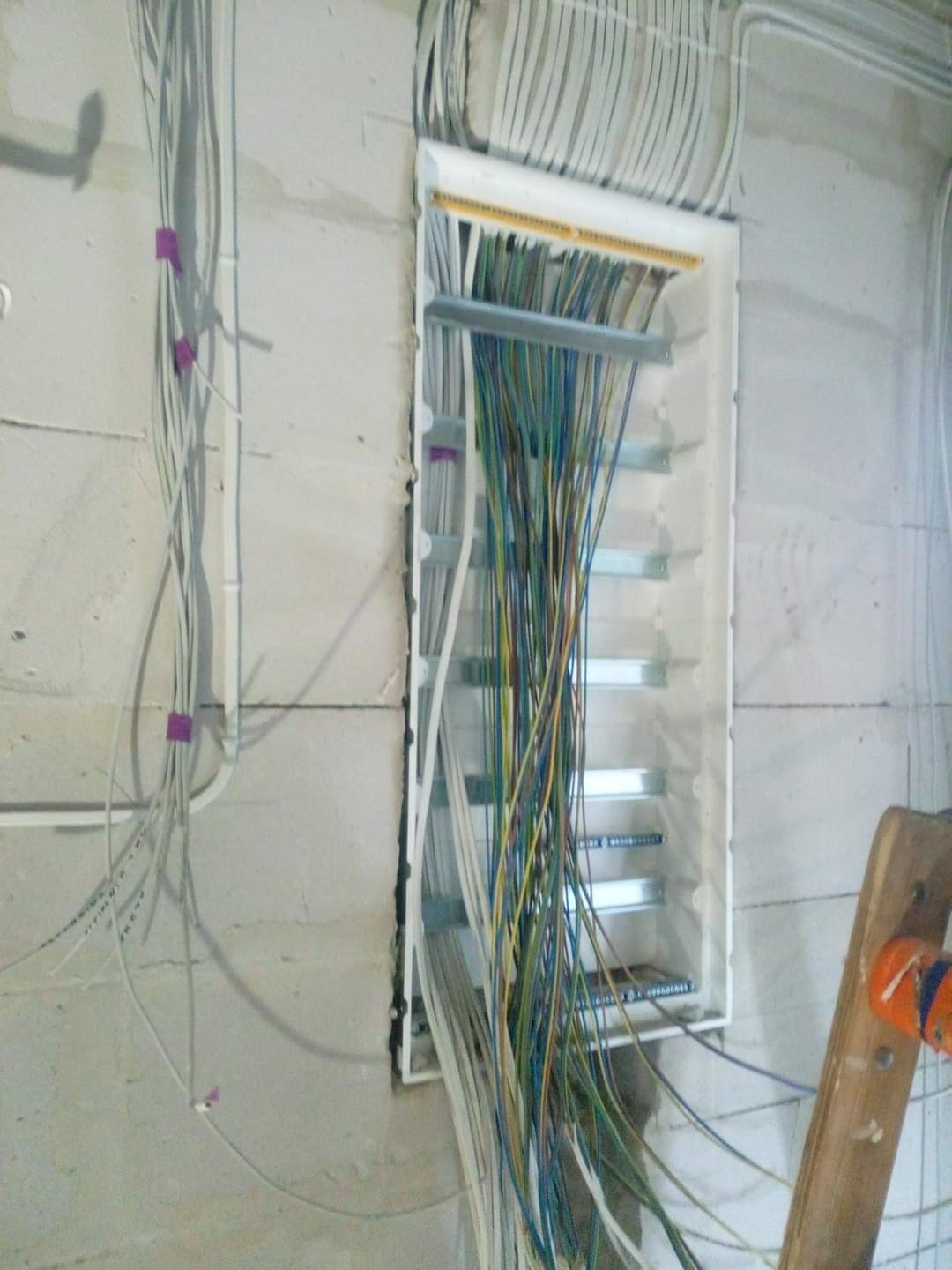Rozdzielnia elektryczna w trakcie montażu w surowym wnętrzu, widoczne przewody w różnych kolorach, częściowo ułożone w korytkach, ściany z betonu komórkowego, fragment drabiny malarskiej z prawej...