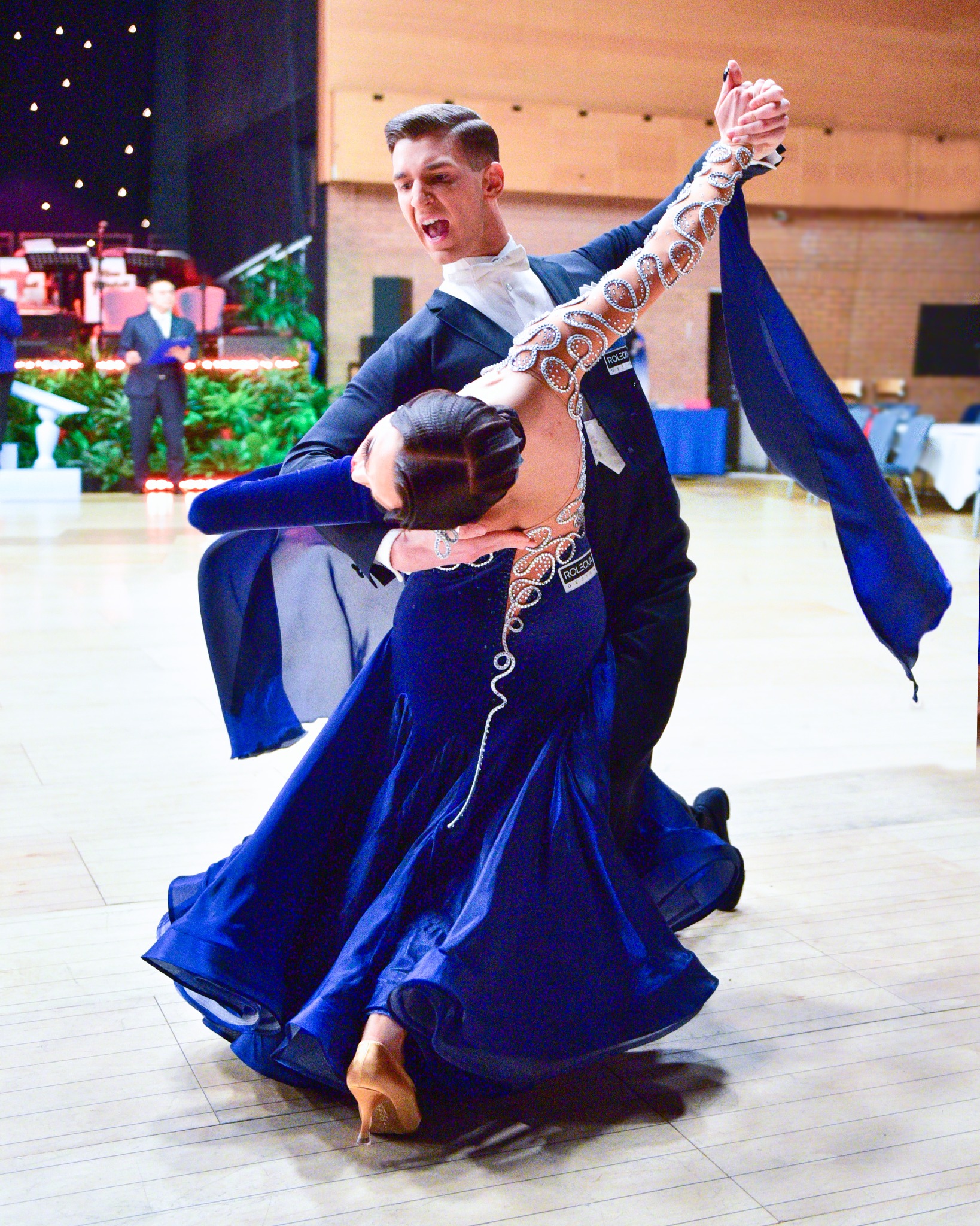 Para tancerzy w dynamicznej pozie na parkiecie, on w czarnym smokingu, ona w granatowej sukni zdobionej kryształkami, w tle dekoracje sceniczne.